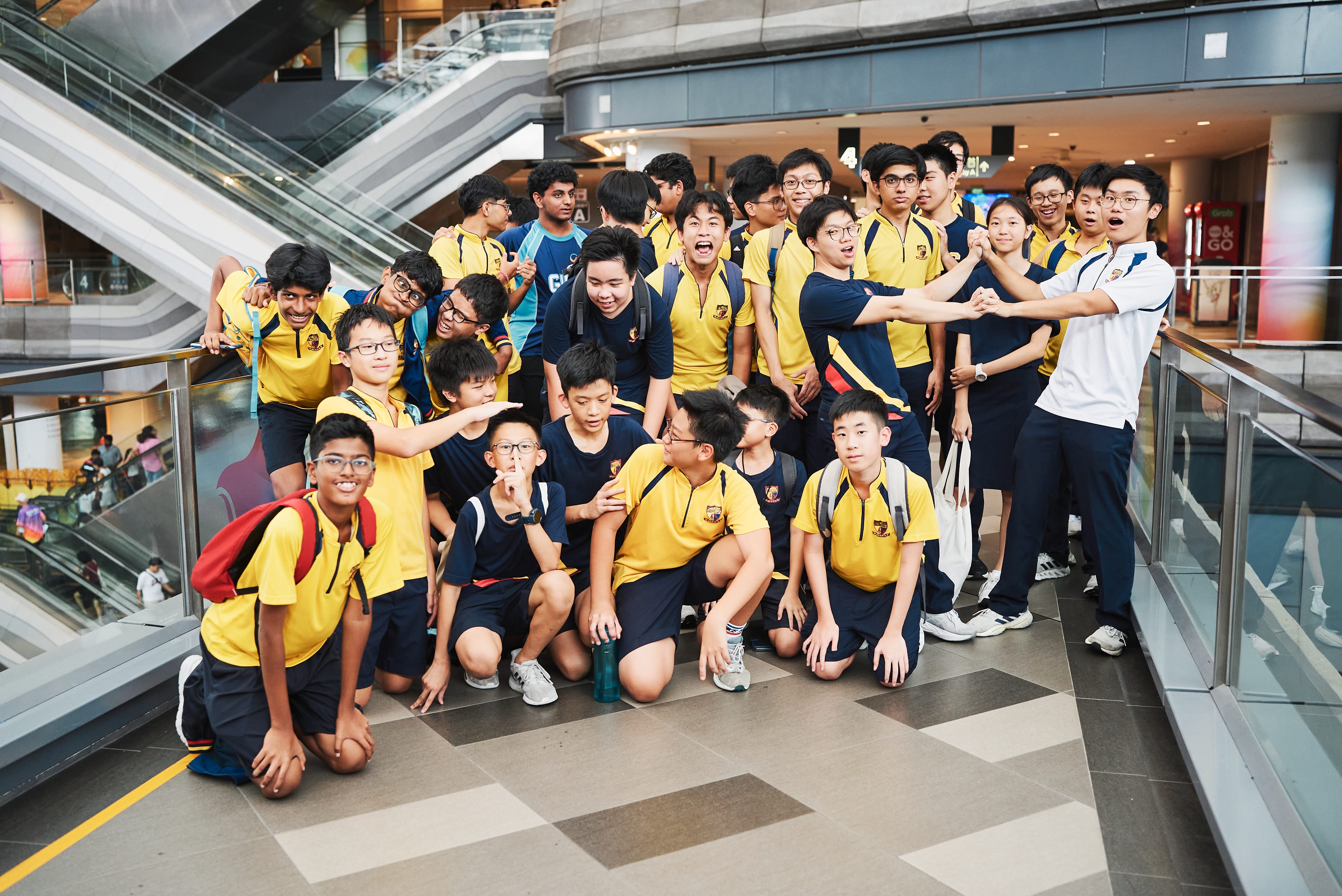 Group of students in yellow and navy uniforms posing and laughing in a mall near escalators