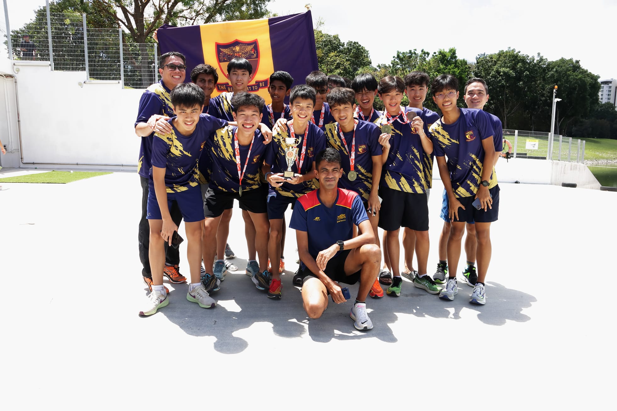 A school sports team in purple and gold jerseys poses with medals, a trophy, and a purple school flag outdoors.