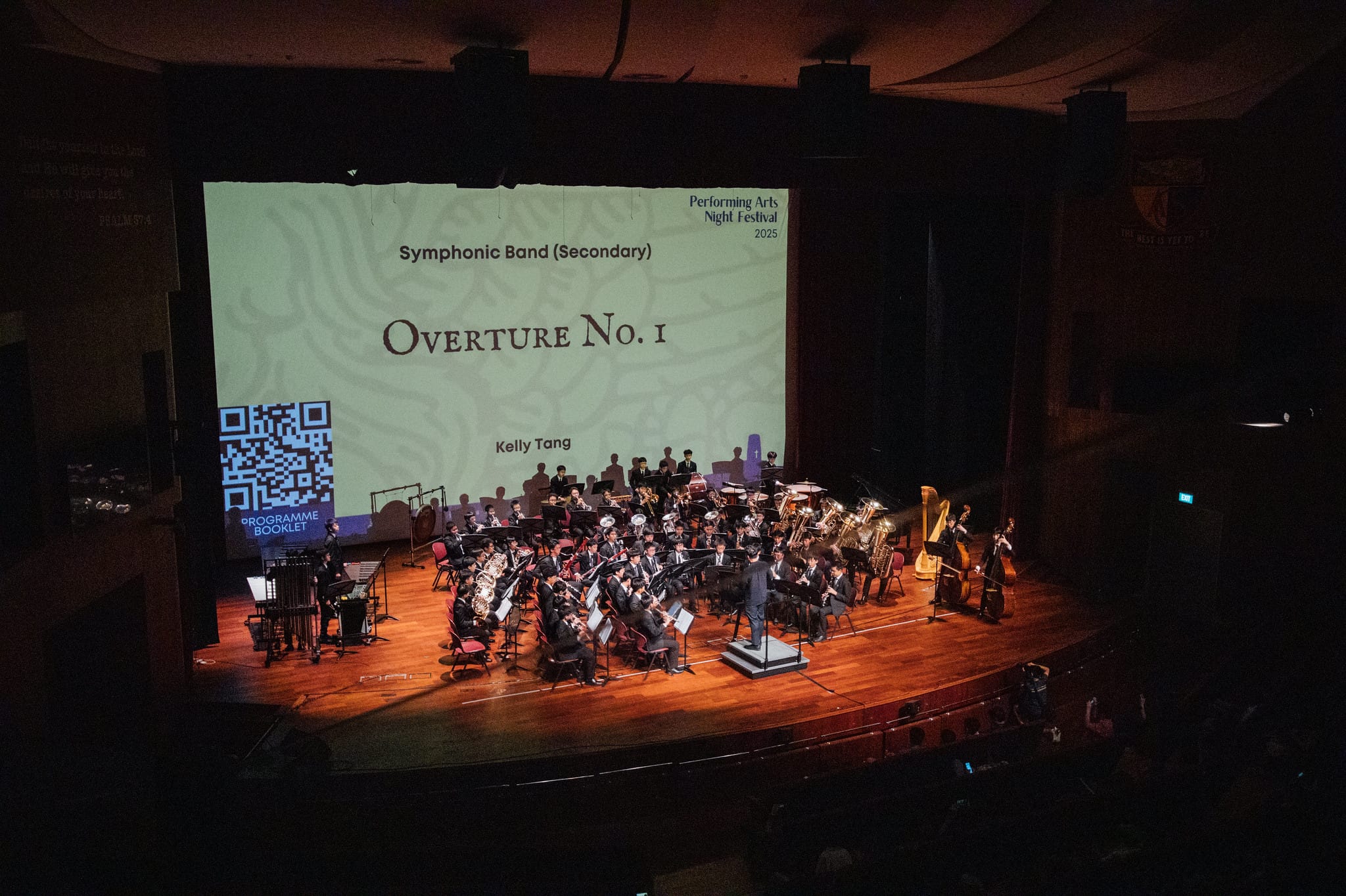 A symphonic band performs on a lit stage with a projected screen reading Overture No. 1 at a Performing Arts Night Festival.