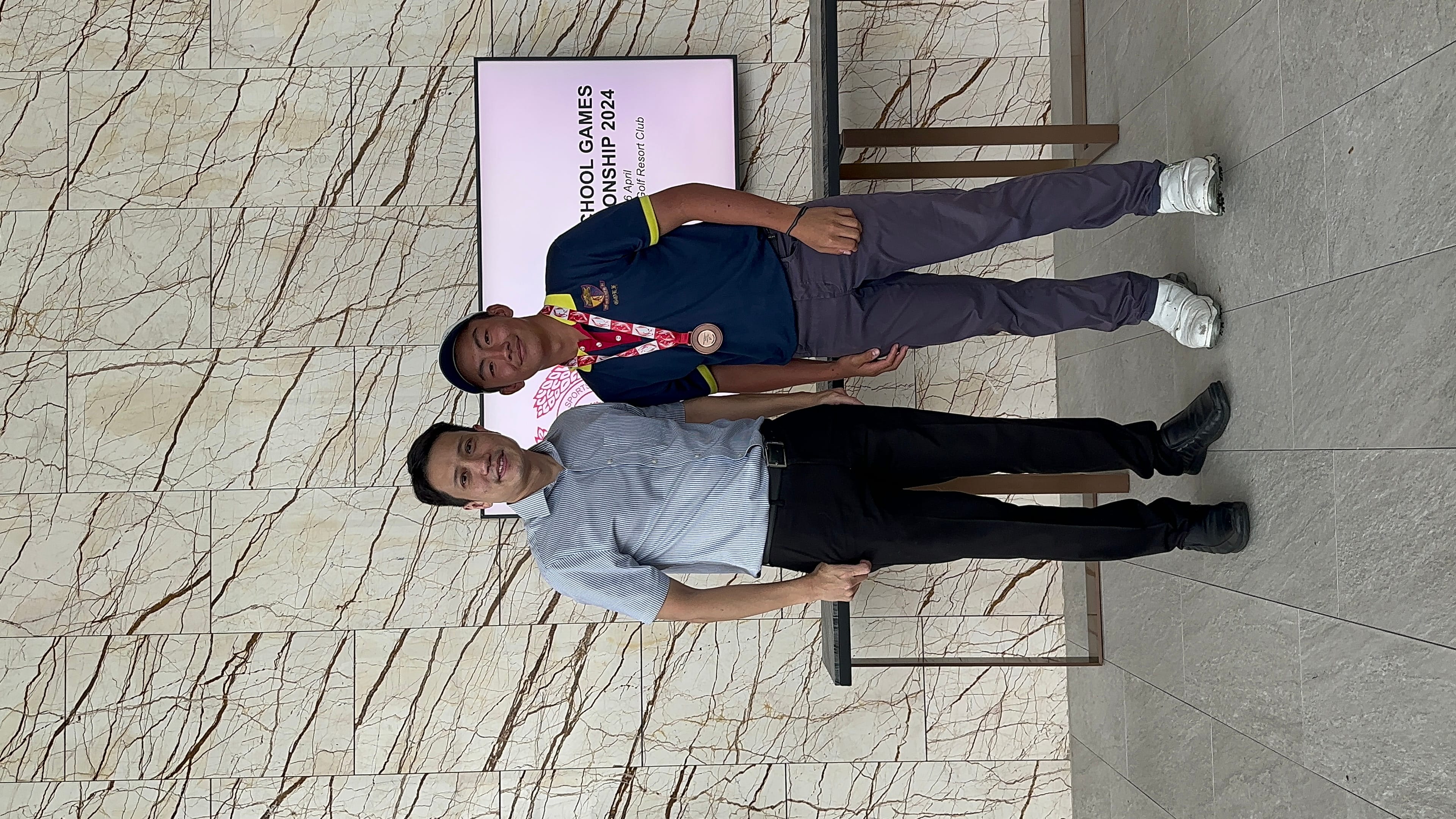 Two men pose in front of a School Games Championship 2024 sign, one wearing a bronze medal and blue cap.