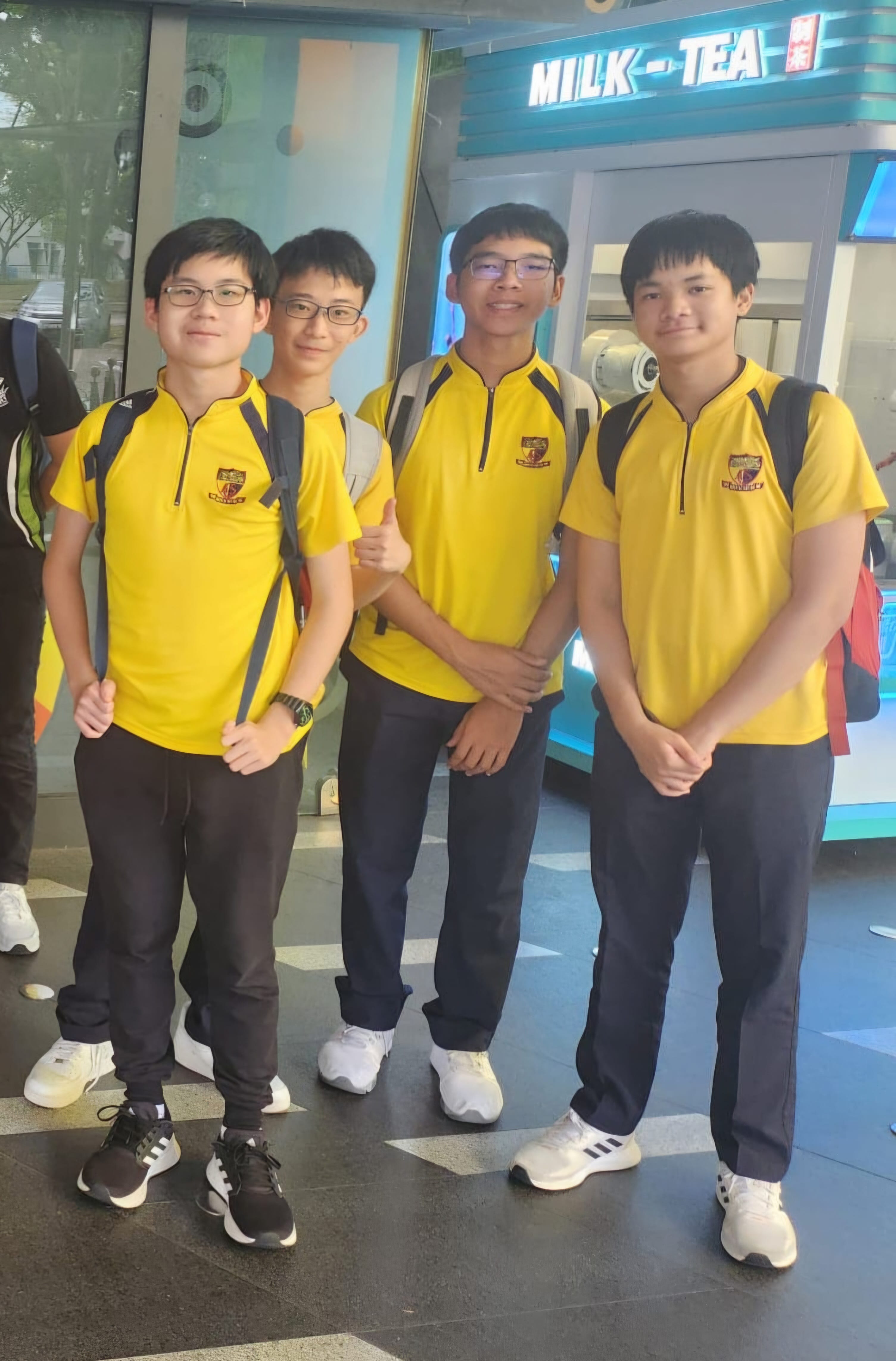 Four smiling students in yellow school uniform pose in front of a milk tea shop. They appear cheerful and are standing together closely.