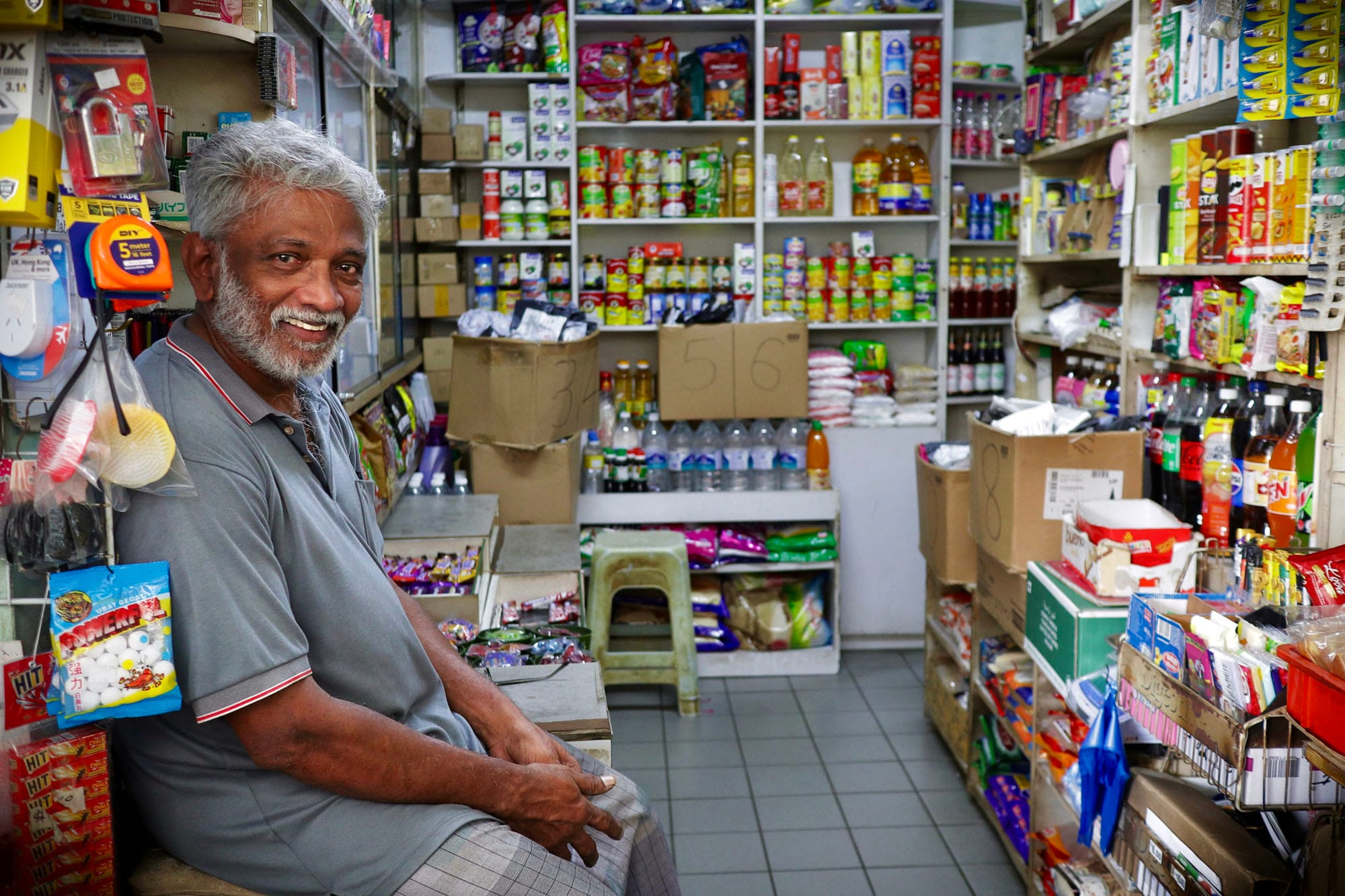 Smiling older man with gray hair sitting inside a small shop packed with shelves of goods and packaged products.