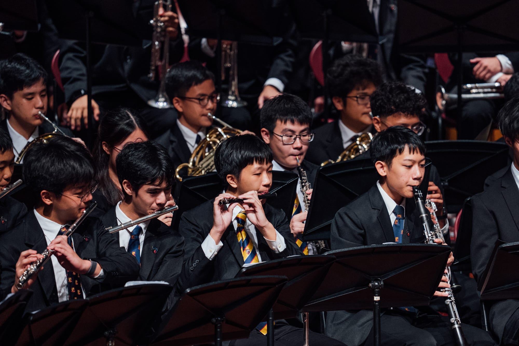 Young students in dark uniforms play flutes, clarinets, and brass instruments in a school orchestra concert.