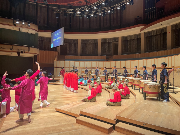 ACS(I) CPAC members perform a combined multicultural showcase on the stage of a professional concert hall, with kompang players in red and pink costumes seated on tiered steps in the foreground and festival drummers in navy uniforms standing behind them. A screen above the stage displays the concert title "Symphony of Cultures."