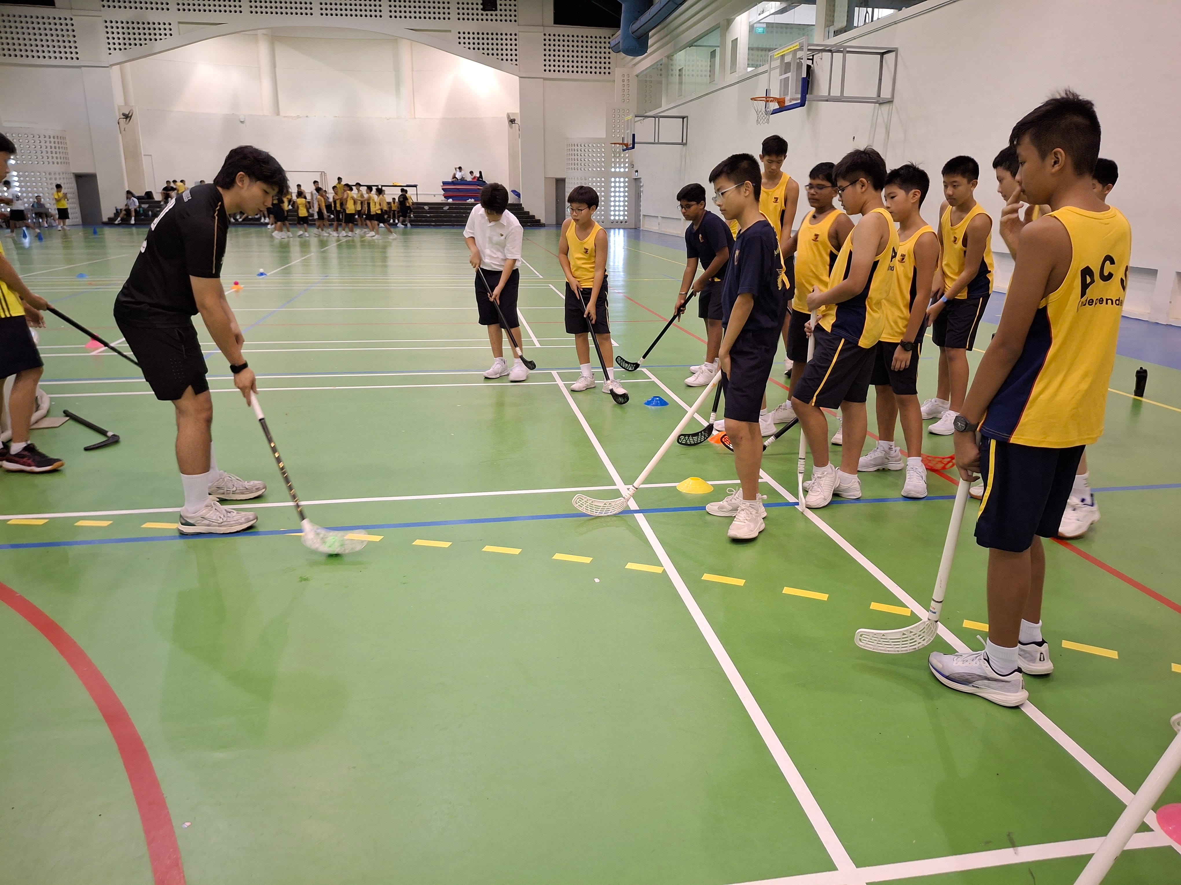 Students in yellow jerseys holding floorball sticks watch a coach demonstrate a drill on an indoor sports court.