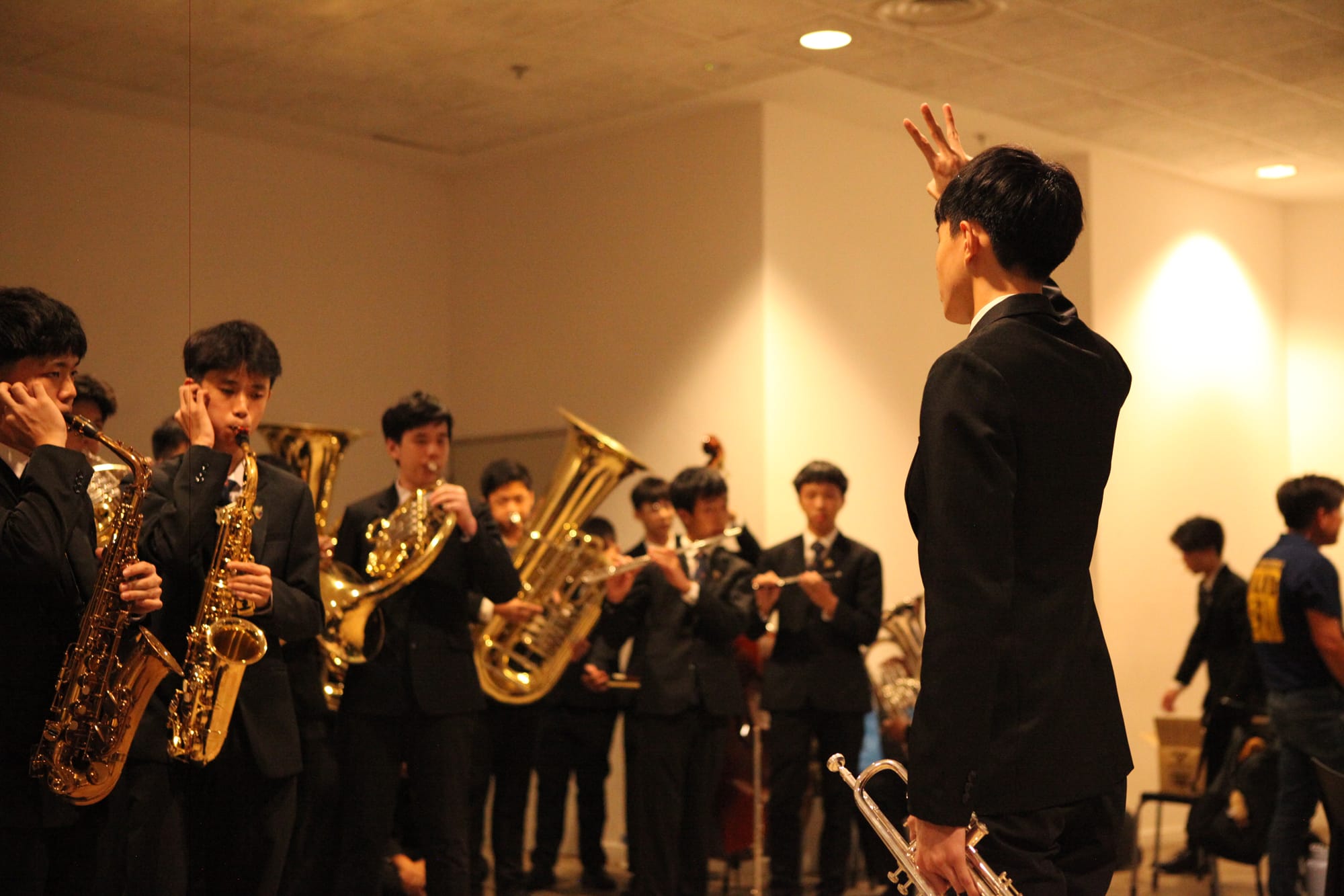 A conductor in a black suit raises his hand directing a youth brass band playing saxophones and tubas indoors.