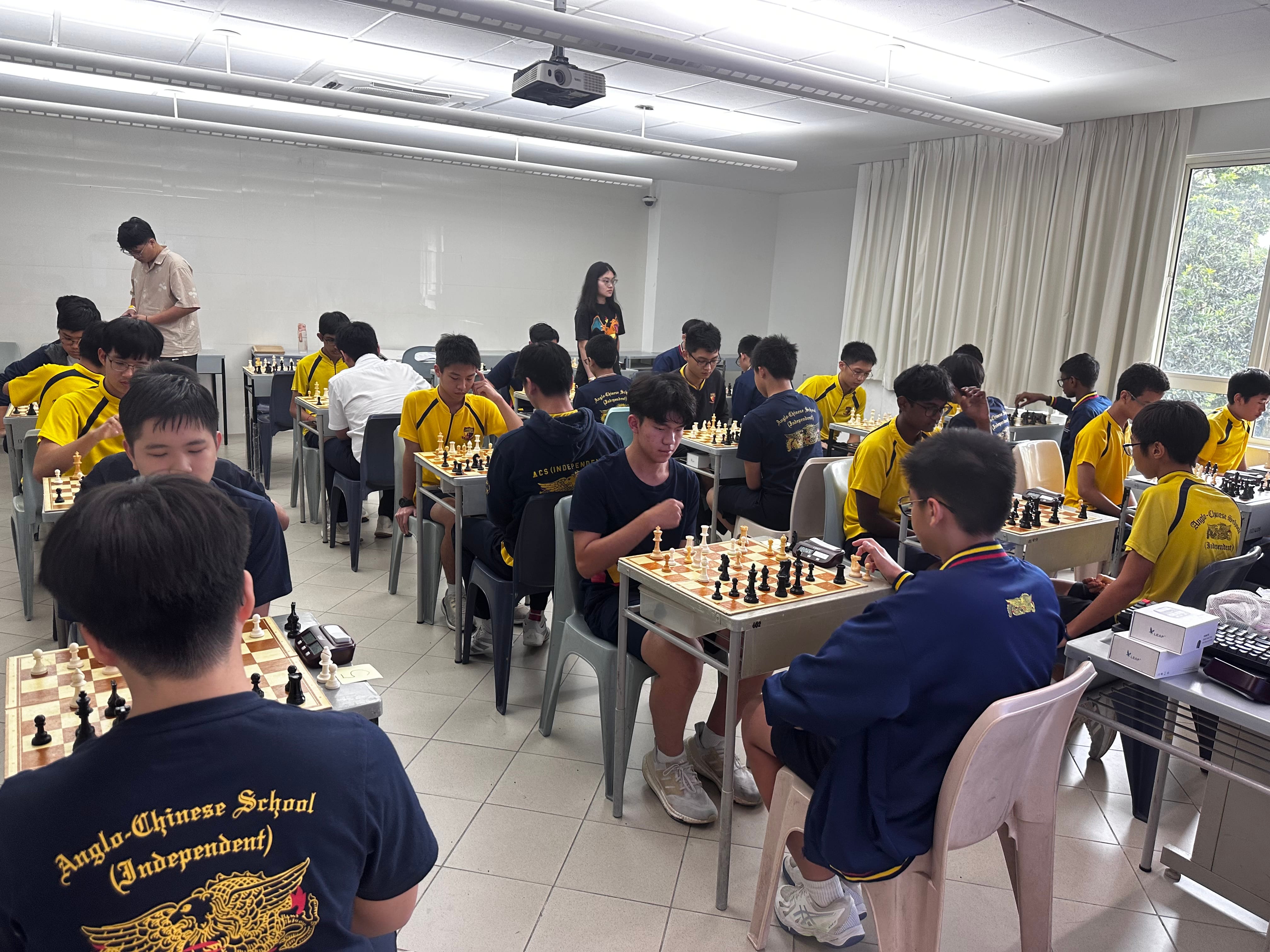 Students in yellow and navy uniforms play chess at multiple tables in a bright classroom during a tournament.