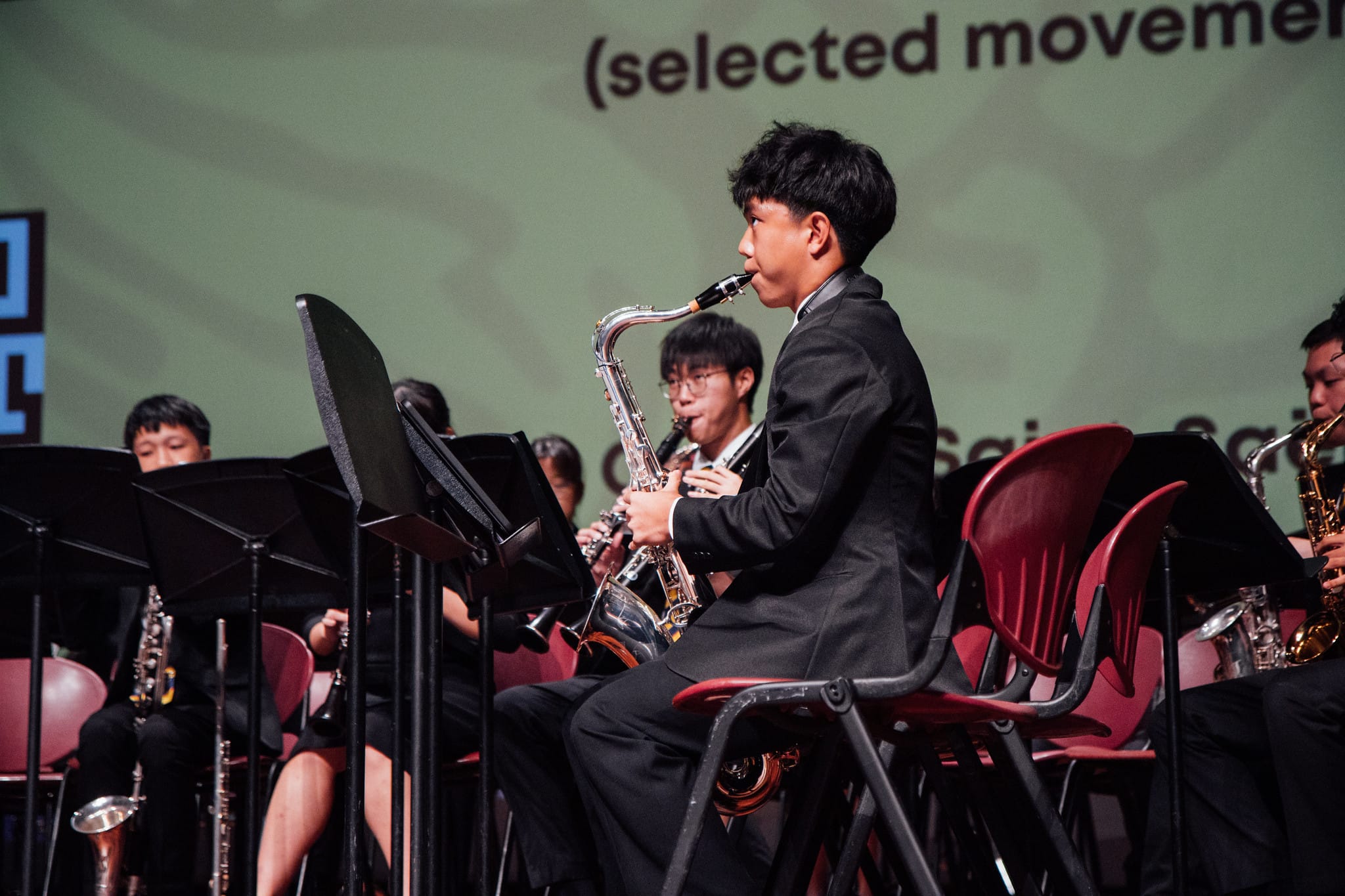Young musicians in black suits sit on red chairs playing saxophones on stage with music stands and a green backdrop.
