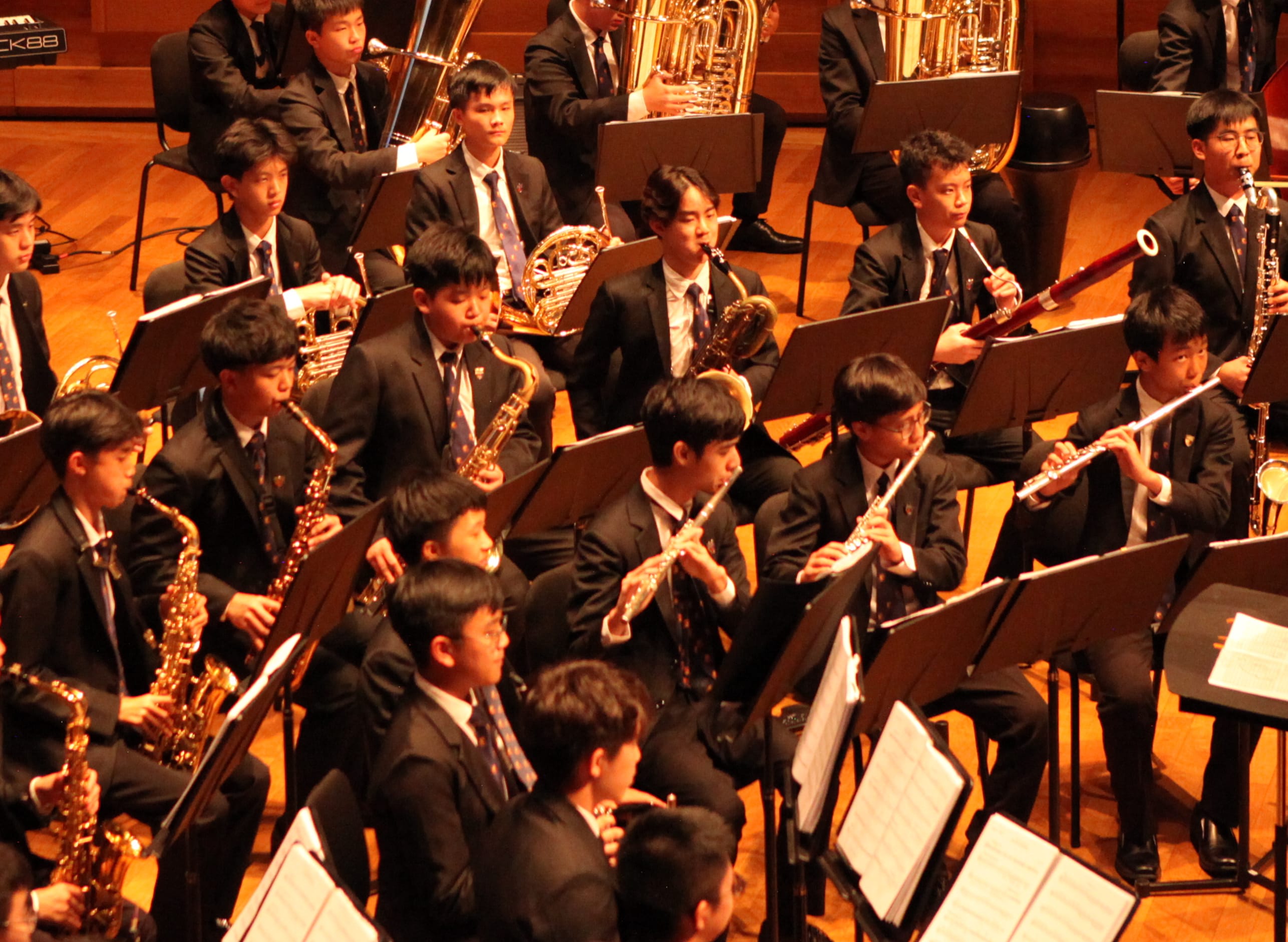 Young students in black suits perform in a large wind band, playing saxophones, flutes, French horns, and tubas on stage.