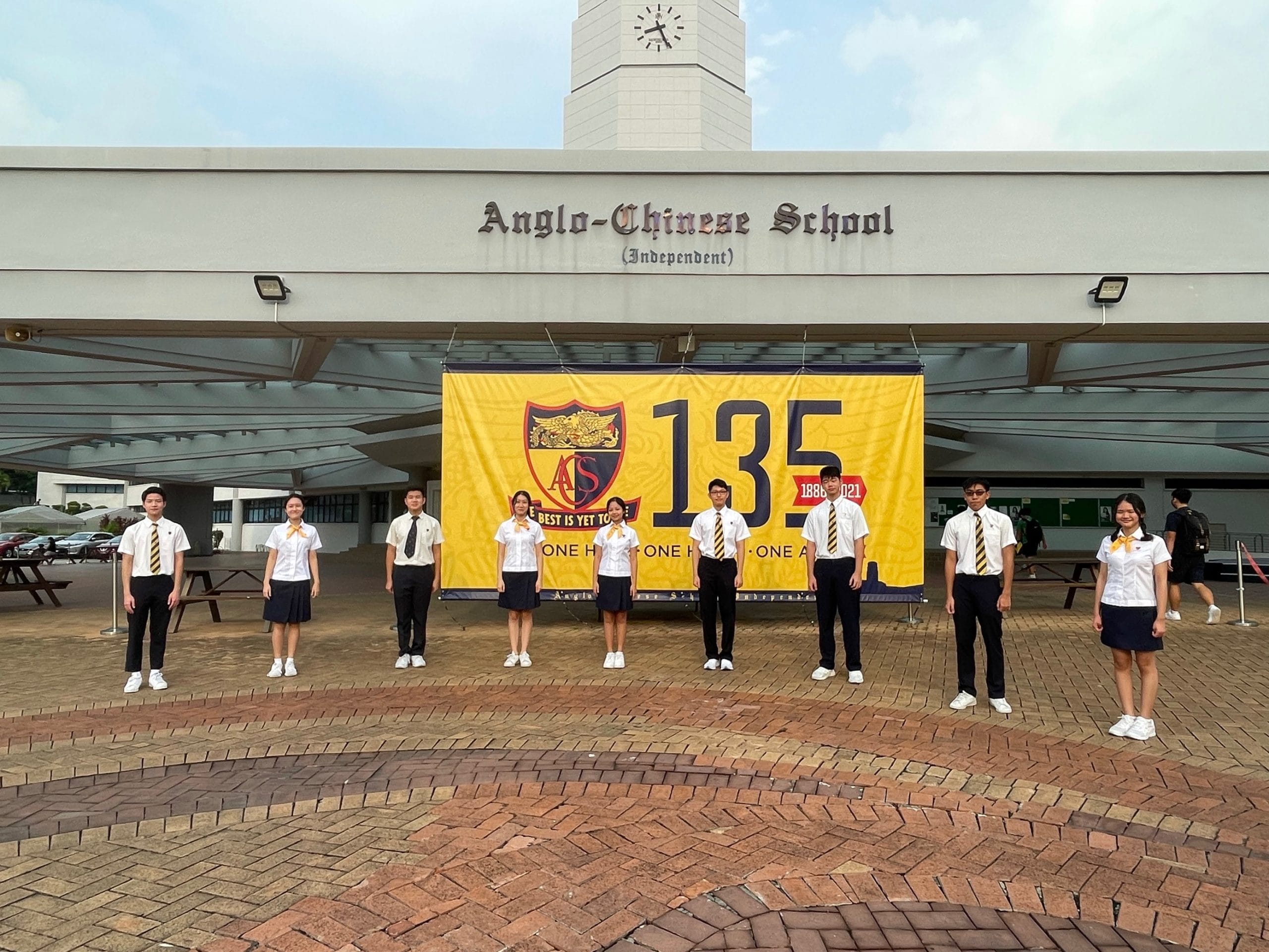 Eight uniformed students stand before Anglo-Chinese School (Independent) building with a yellow 135th anniversary banner.