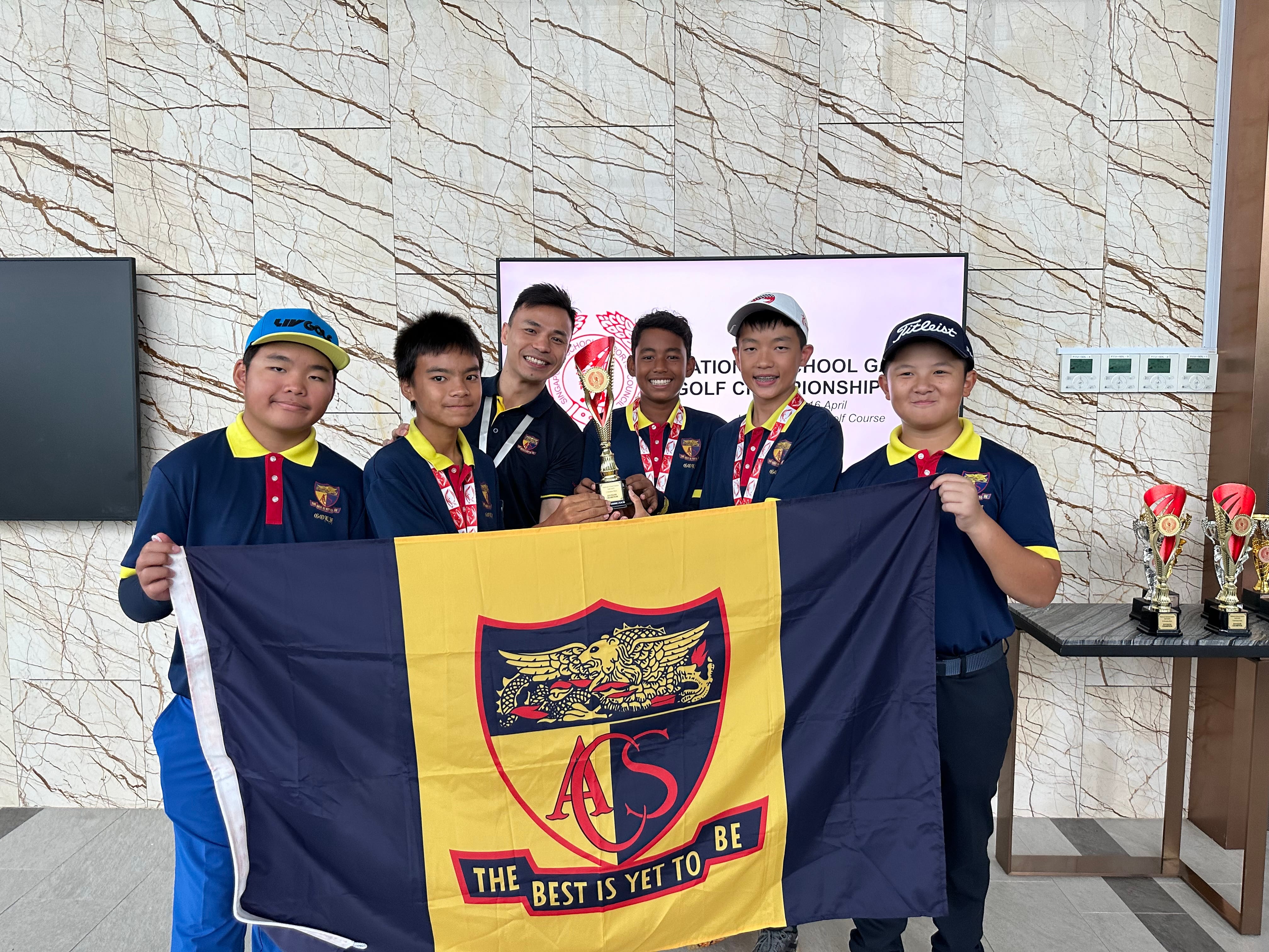 Five boys and a man in matching polos hold a large flag and a trophy; more trophies on table.
