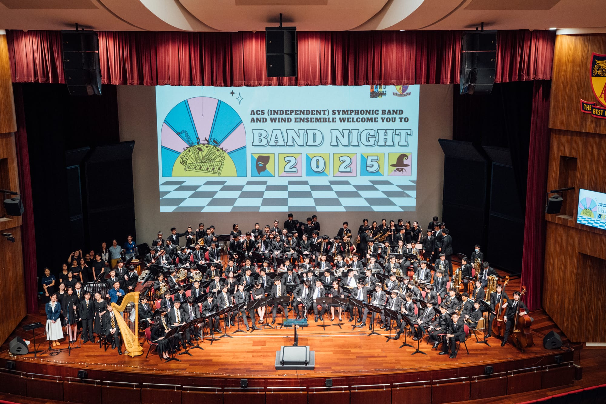 Large school symphonic band on stage in formal attire with a Band Night 2025 banner projected behind them.