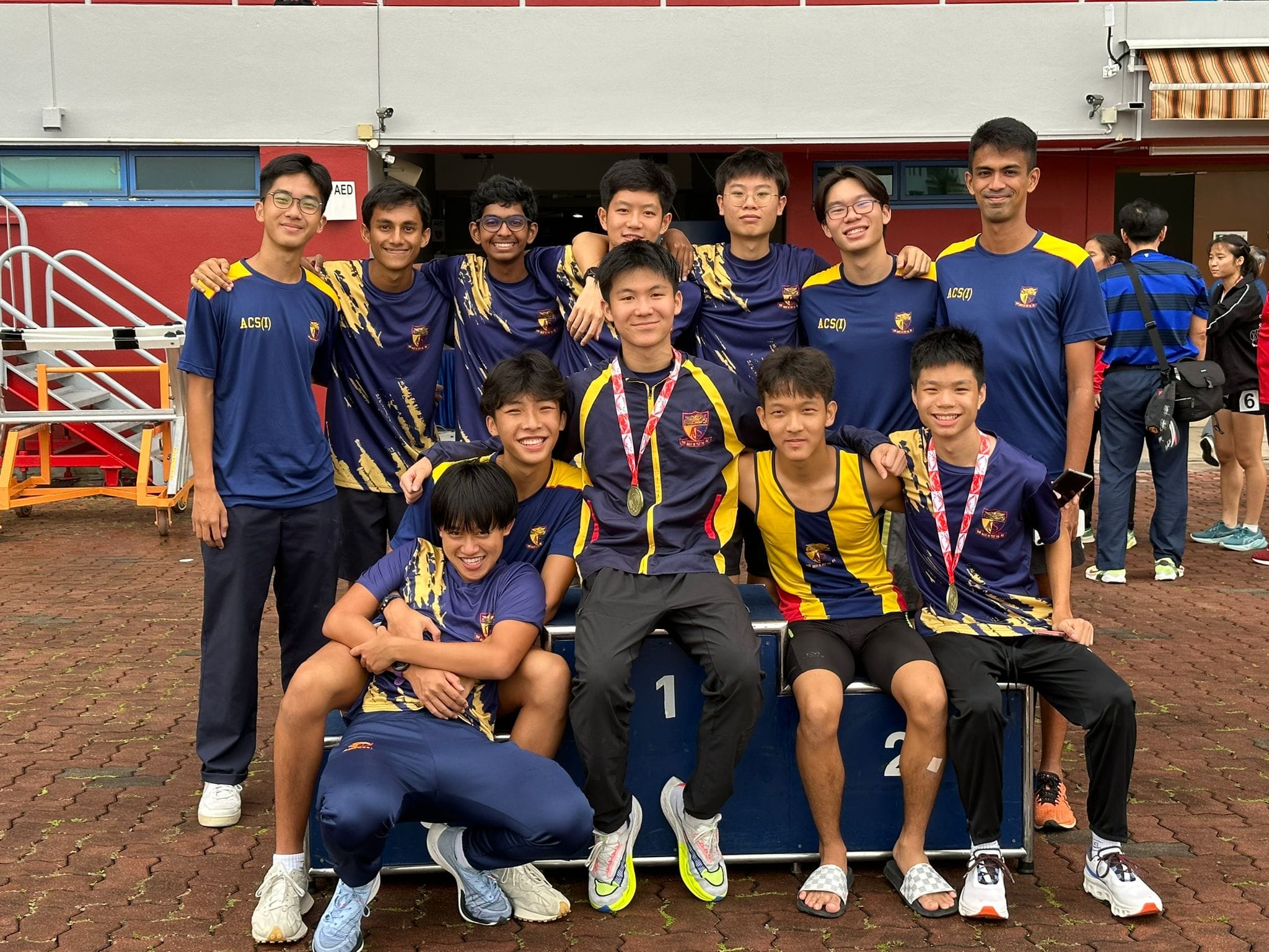 A group of male students in navy and gold ACS(I) uniforms pose together outdoors, some wearing medals, near a podium.