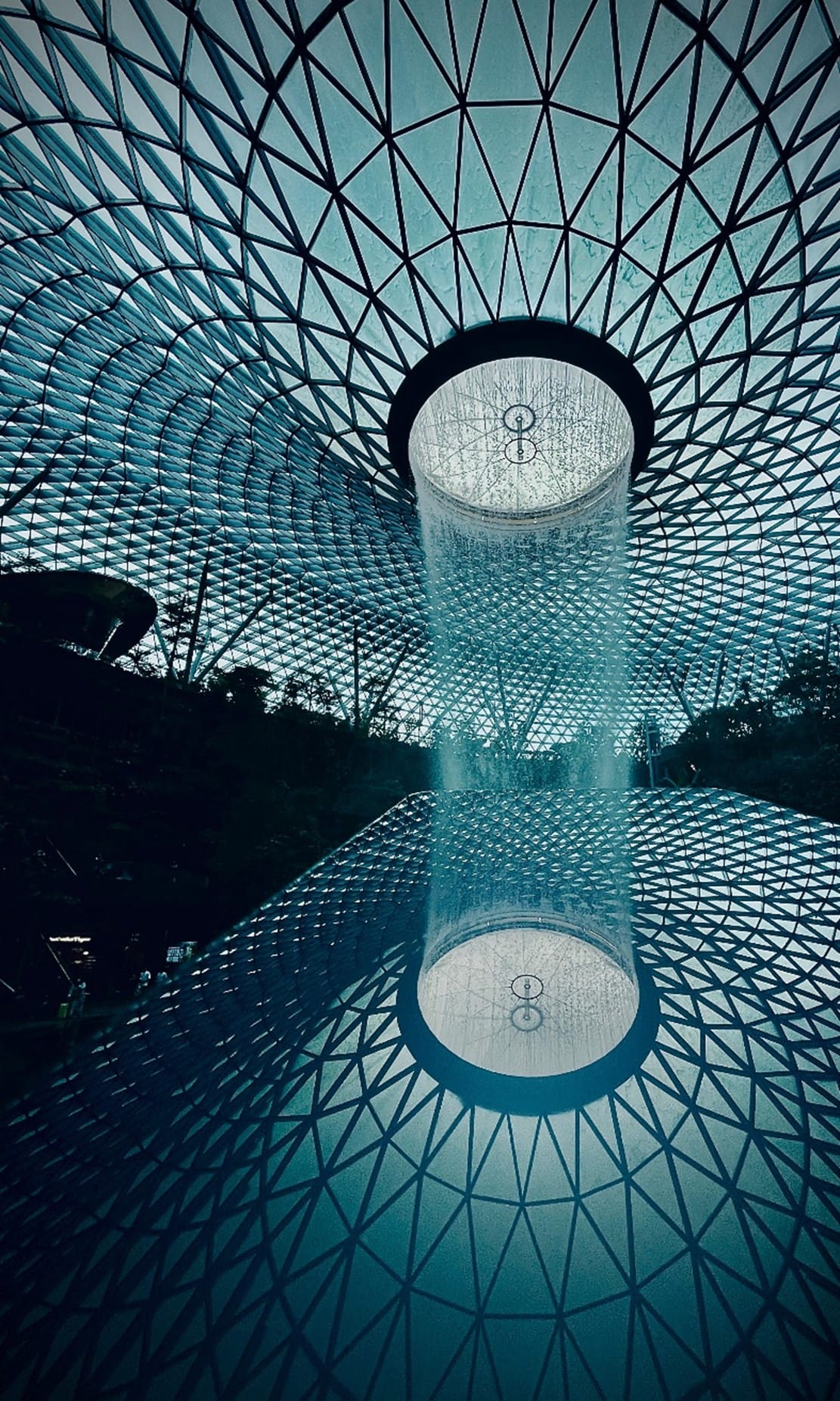 Indoor waterfall descending from a circular opening in a massive geometric glass and steel domed roof, reflected below.