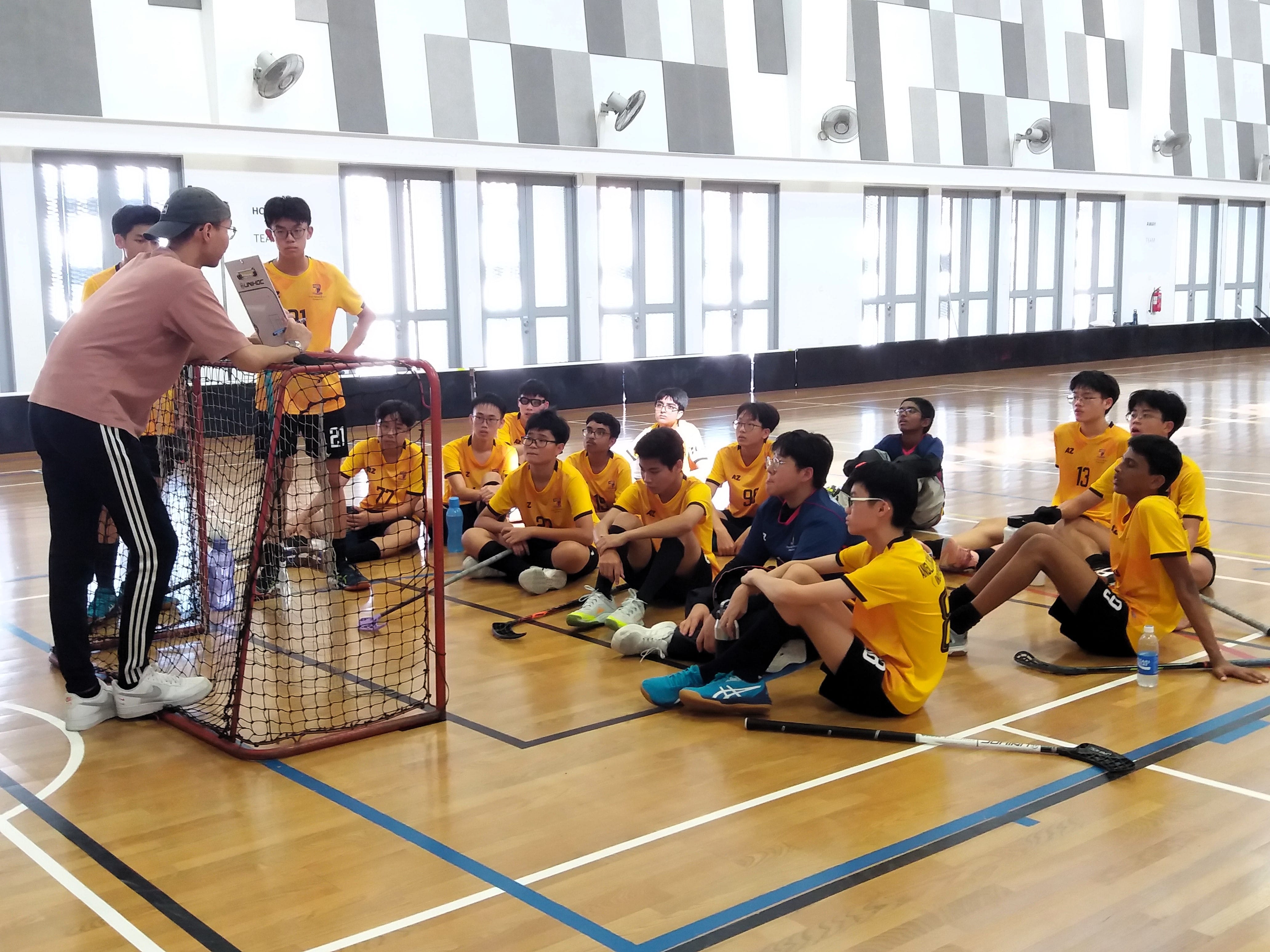 A coach addresses a youth floorball team in yellow jerseys seated on an indoor gym floor near a goal net.