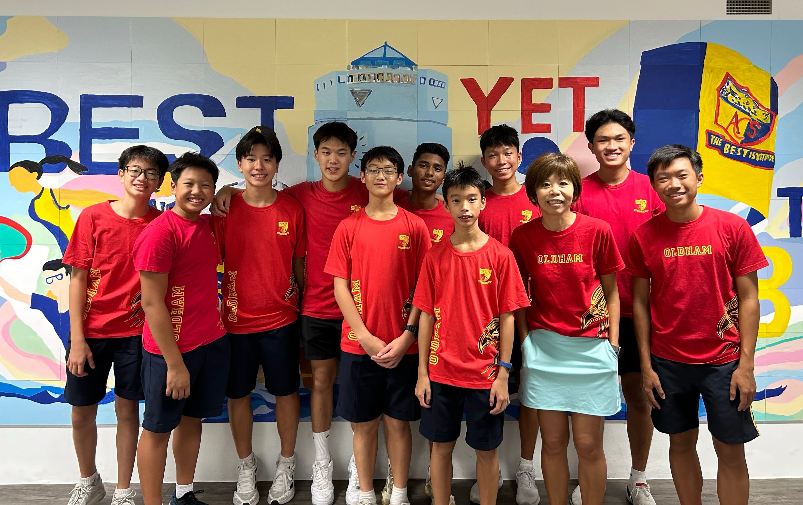 A group of eleven people wearing red Oldham shirts pose smiling in front of a colorful mural reading Best Yet.