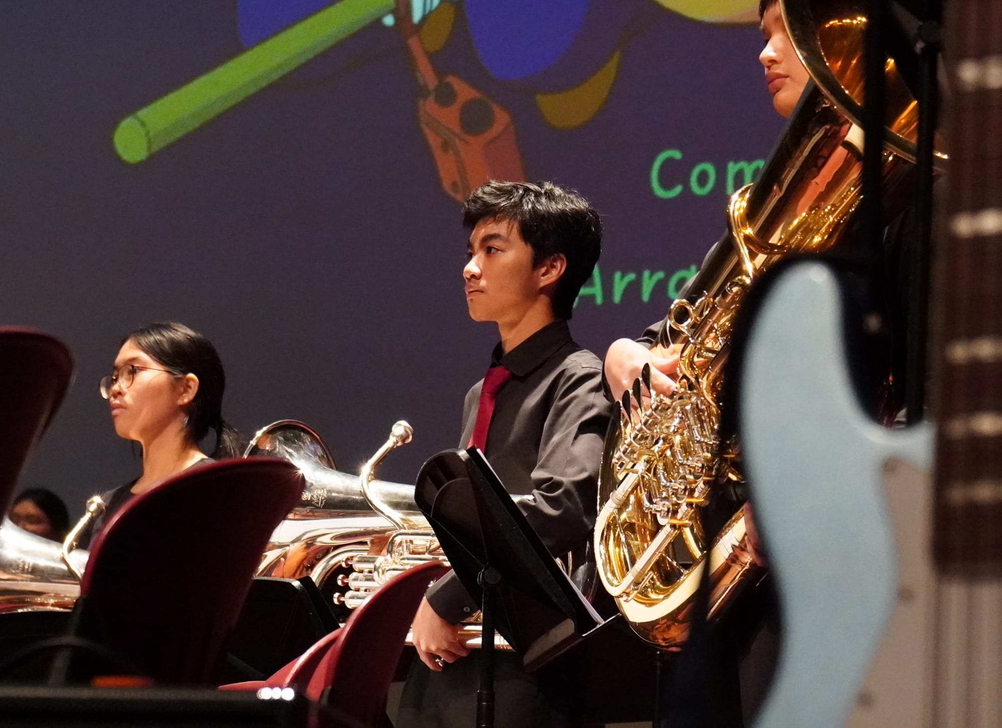 Young musicians in black formal wear hold brass instruments on stage before a colorful projected backdrop.
