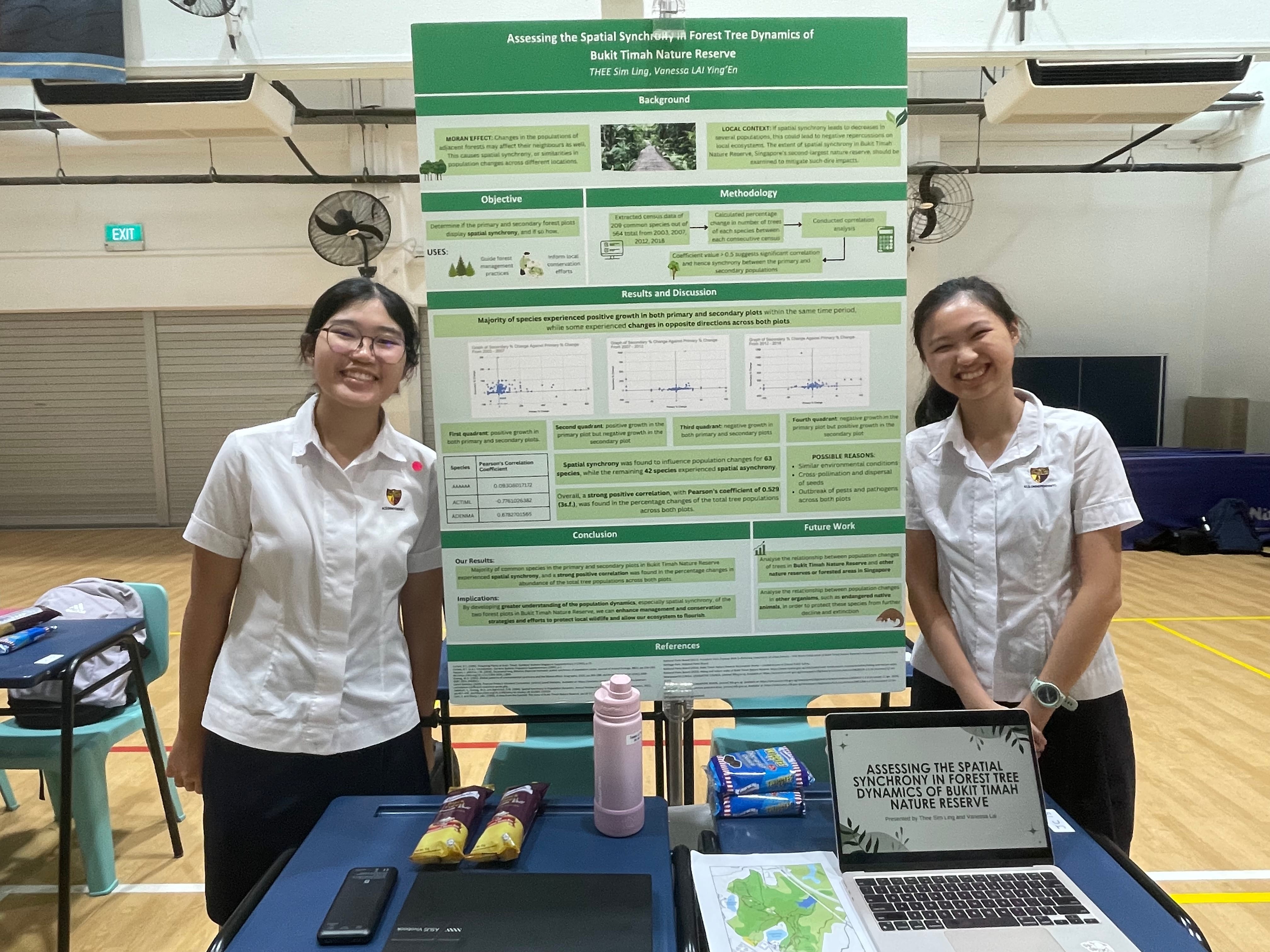 Two students in school uniform smile beside a green and white science project poster in a bright, spacious hall. On the table, the laptop screen reads "Assessing the spatial synchrony in forest tree dynamics of Bukit Timah Nature Reserve".