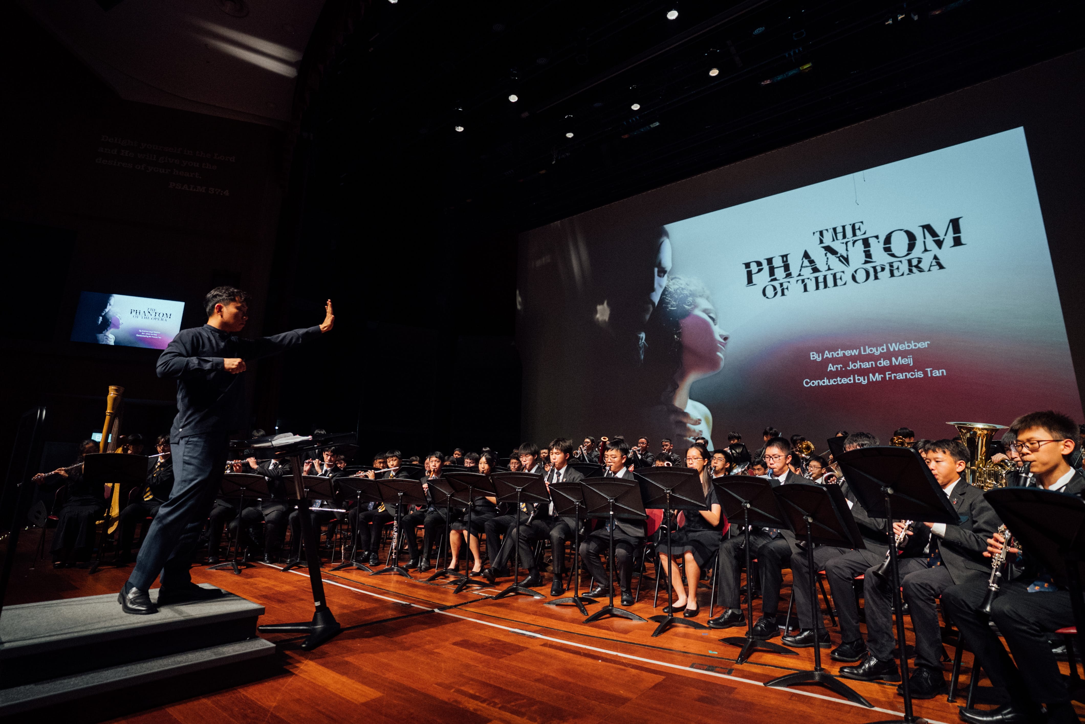 A conductor leads a youth wind orchestra performing Phantom of the Opera, with a large projection screen behind them.