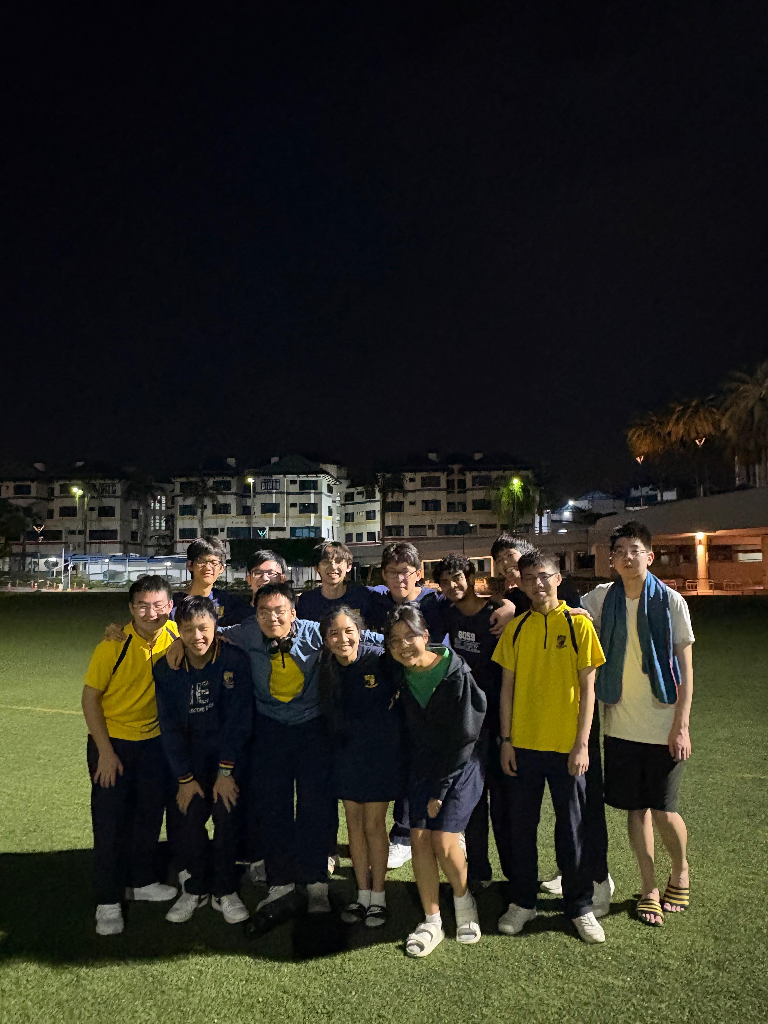 A group of about 15 young people posing together on a green field at night with buildings in the background.