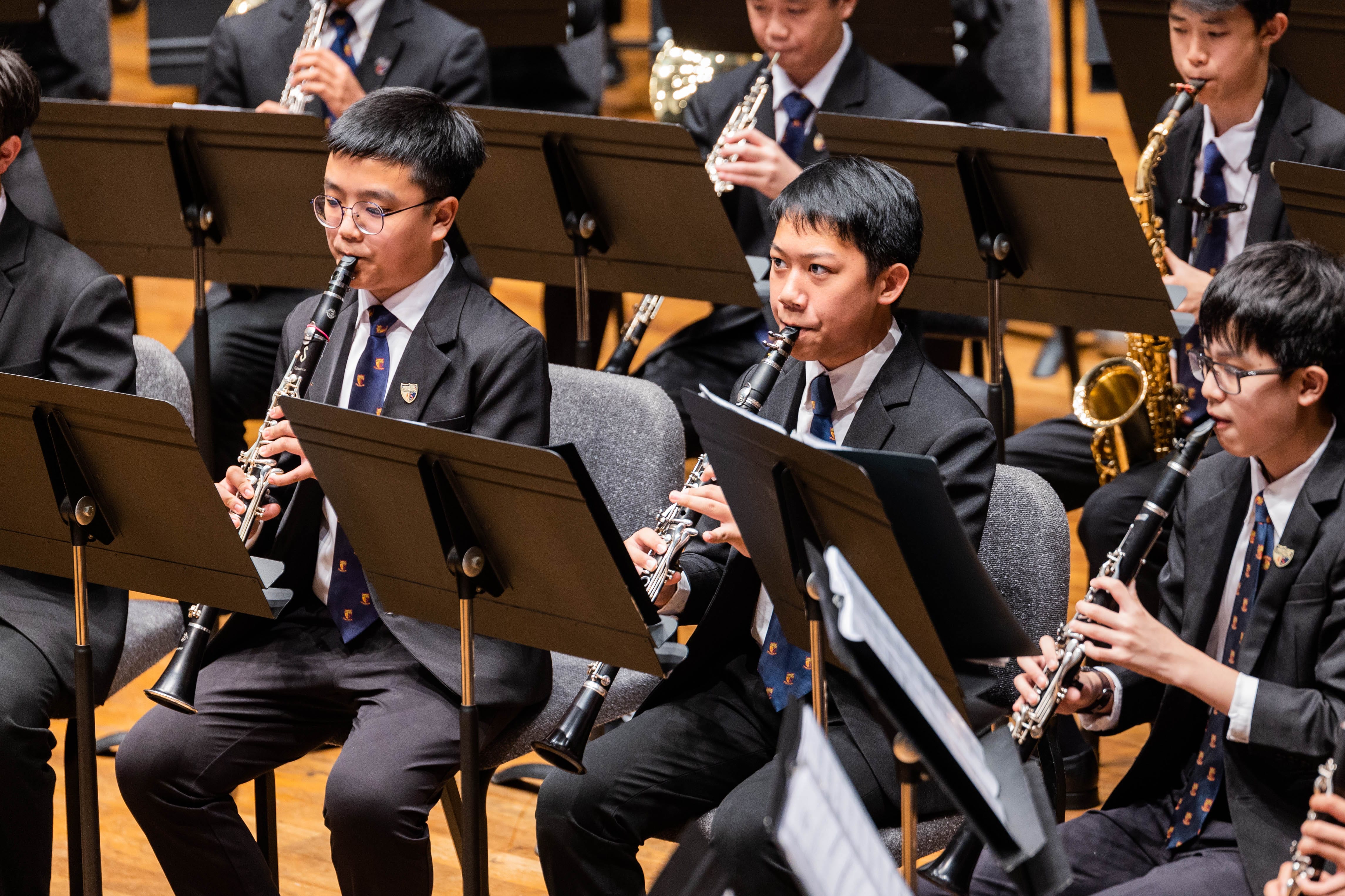 Young musicians in black suits and ties play clarinets and saxophones during a formal concert performance.