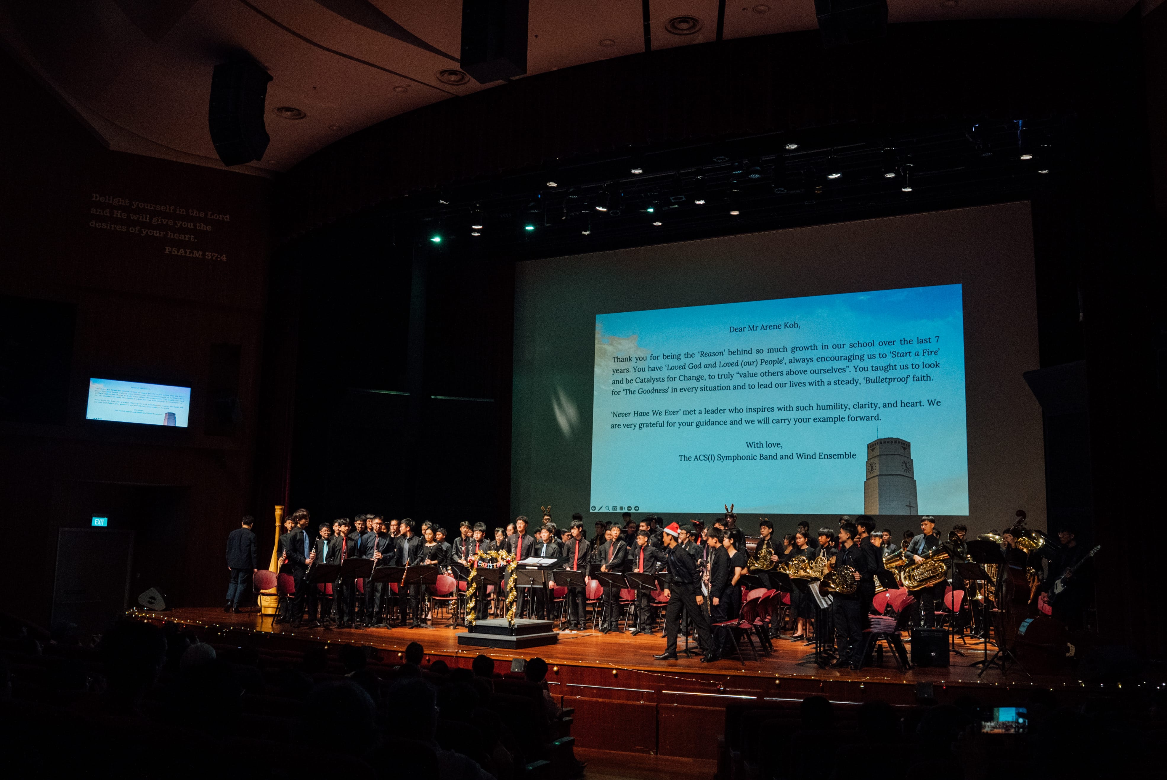 A school band in black attire performs on a dimly lit stage with a large projected thank-you letter on screen behind them.