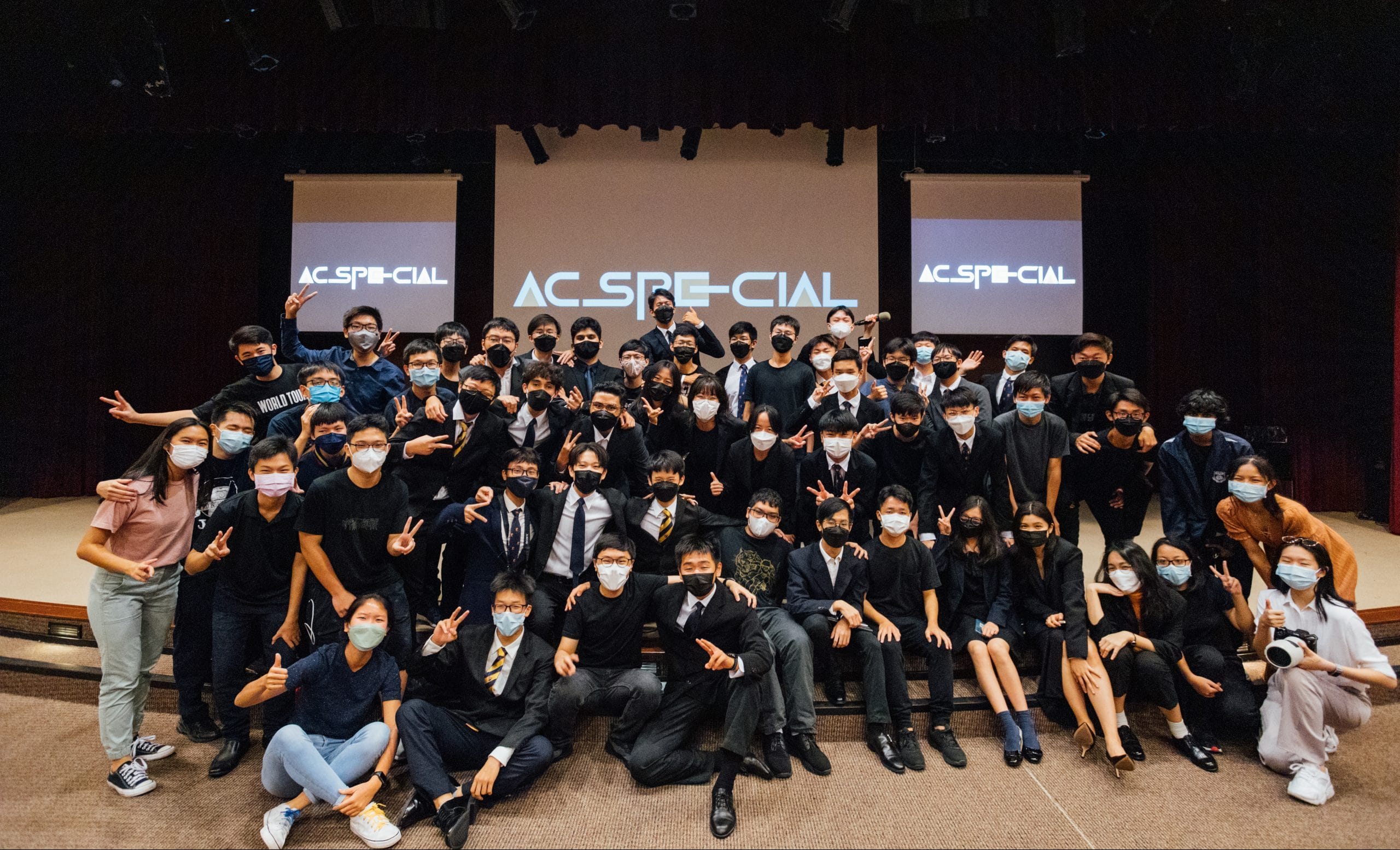 Large group of masked young people posing on a stage in front of screens displaying AC_SPECIAL.