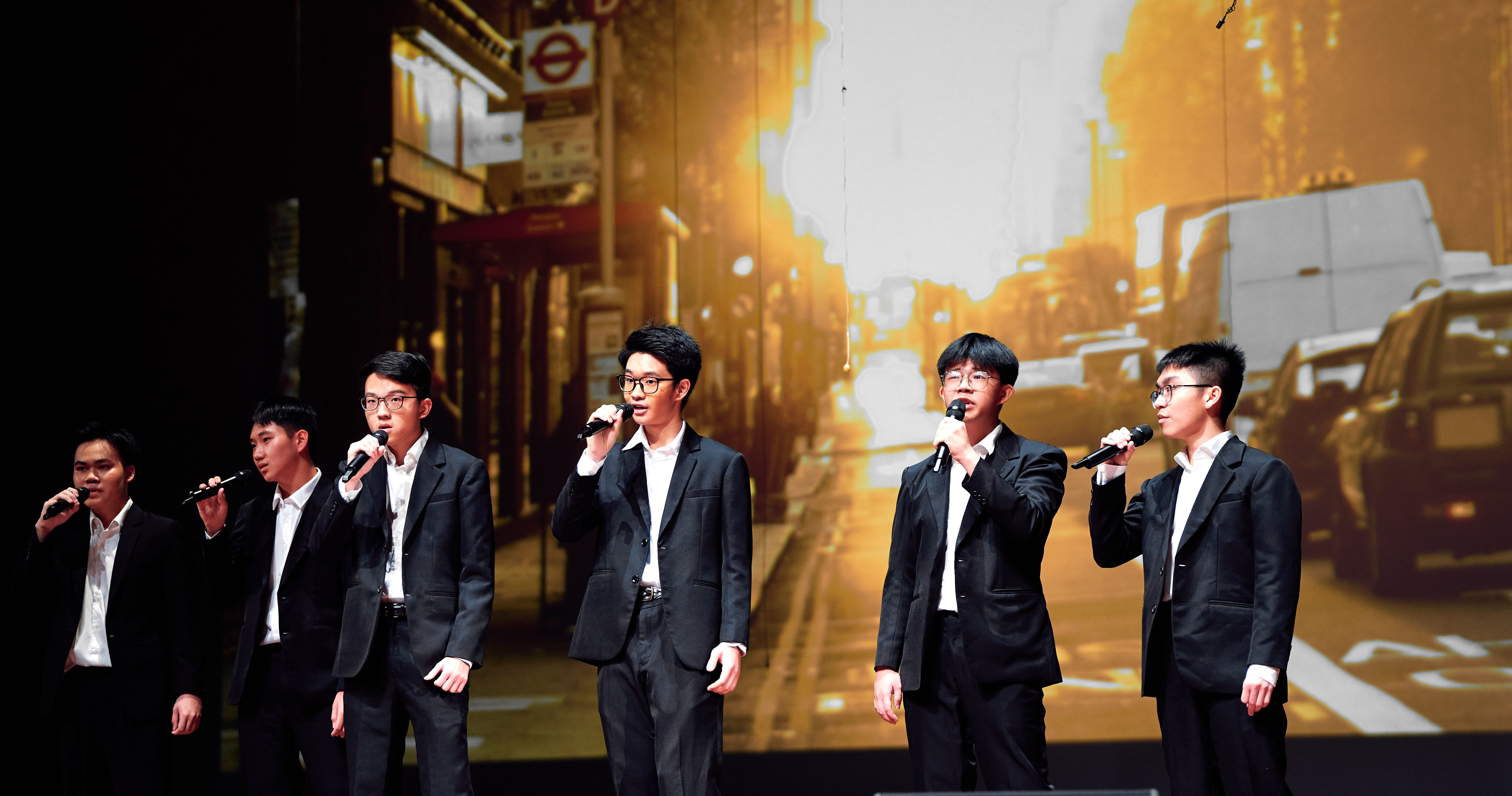 Six young men in black suits sing into microphones on stage before a projected street scene backdrop.