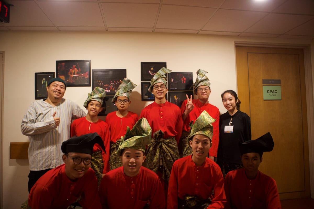 ACS(I) CPAC kompang members in red baju melayu and songkok headgear pose backstage with a teacher and a student conductor outside a dressing room door labelled "CPAC," smiling after a performance at a professional concert venue.