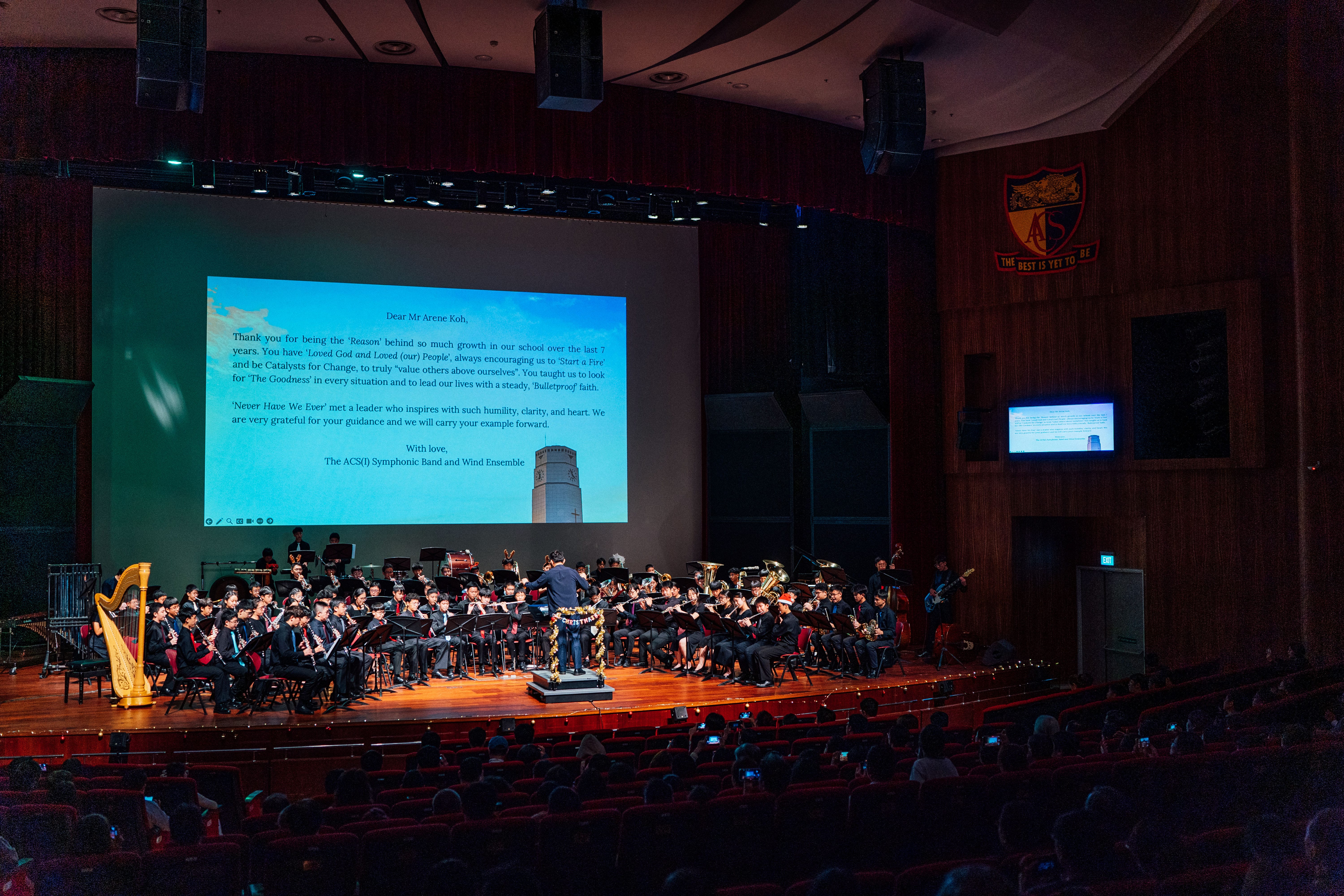 School concert hall with conductor leading band onstage beneath a large screen showing a letter.