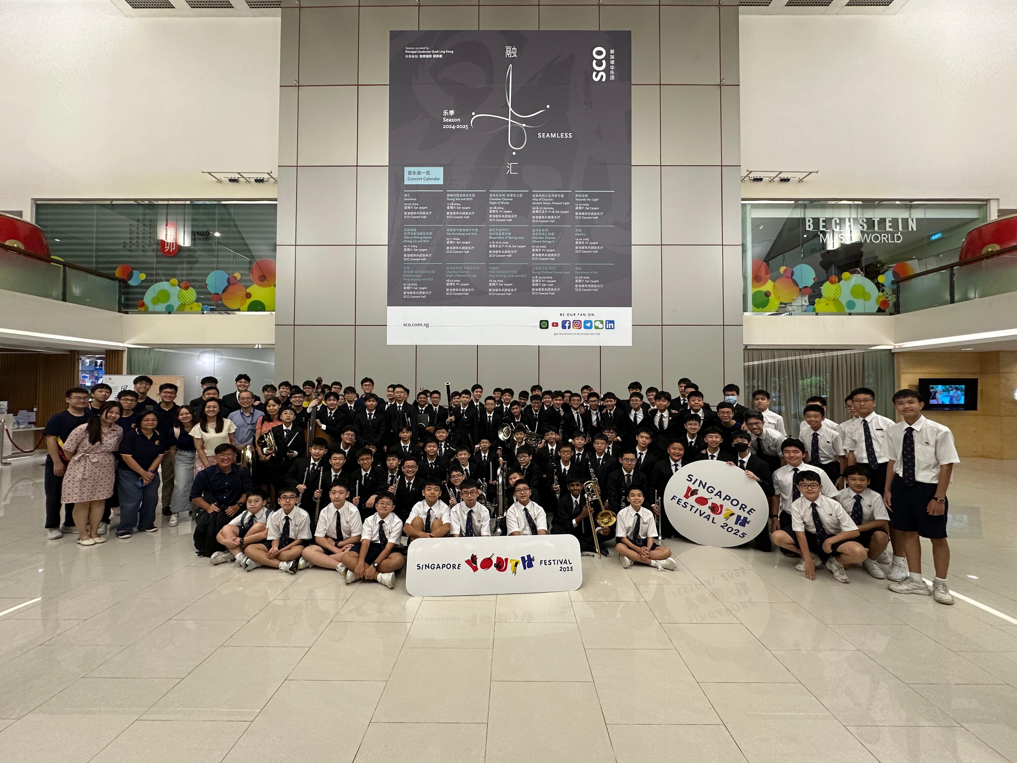 Large group of uniformed youth musicians posing indoors with Singapore Youth Festival 2025 signs and instruments.