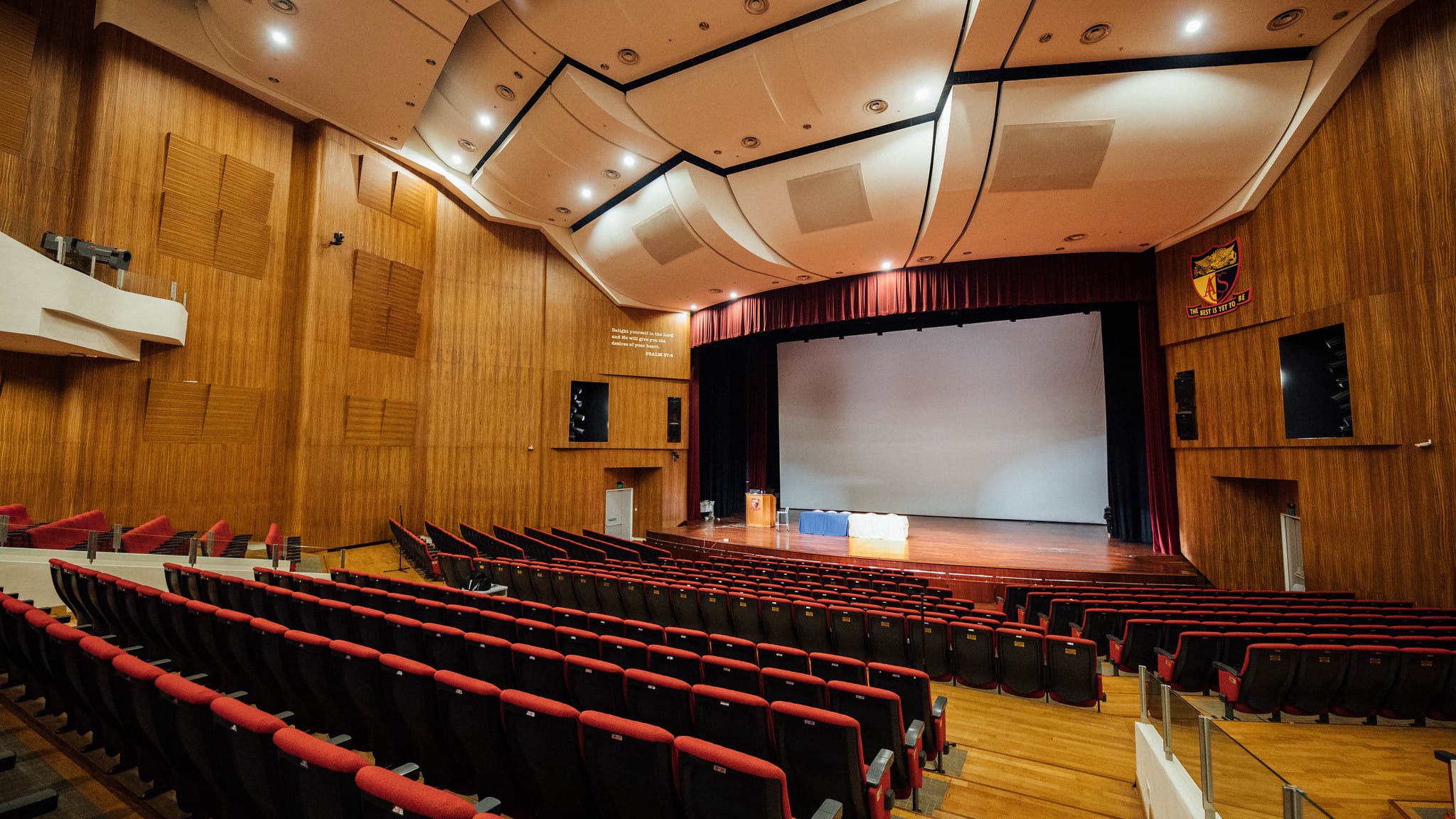 A school auditorium with red seats, a wooden stage, red curtains, and a projected screen showing green slides.