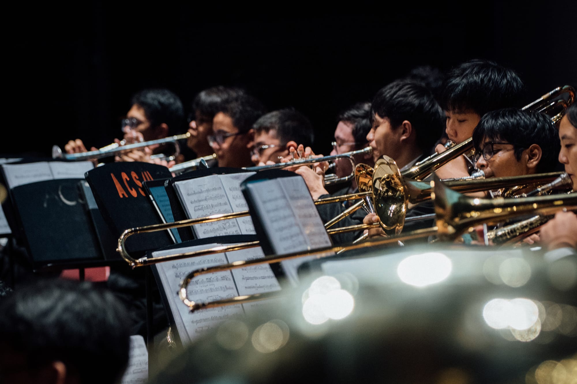 Young musicians play trombones and brass instruments in a row during a concert, reading sheet music on stands.