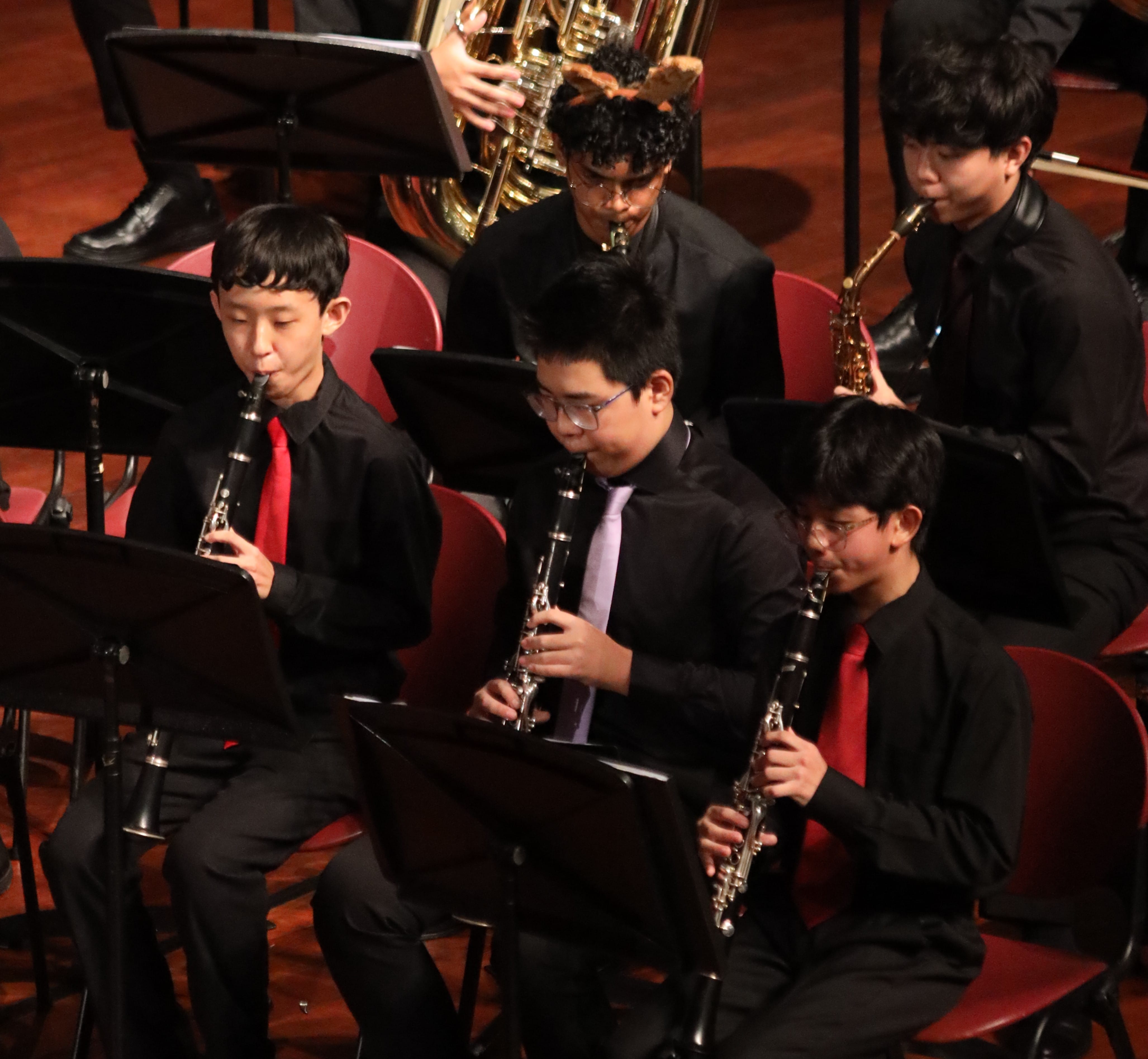 Young students in black attire play clarinets and brass instruments seated in red chairs during a concert performance.