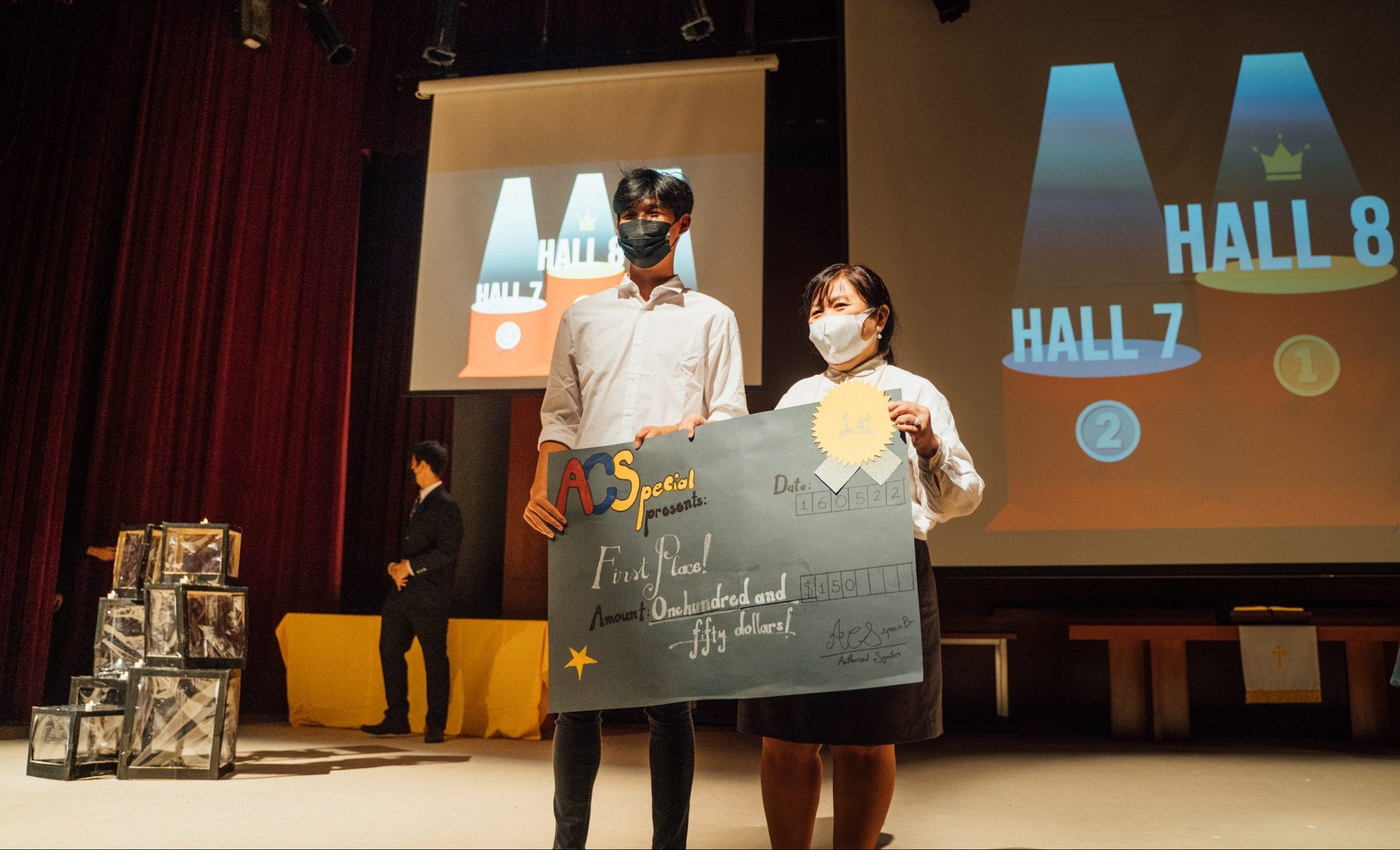 Two masked students on stage hold a large check for first place, one hundred fifty dollars, from ACS Special.