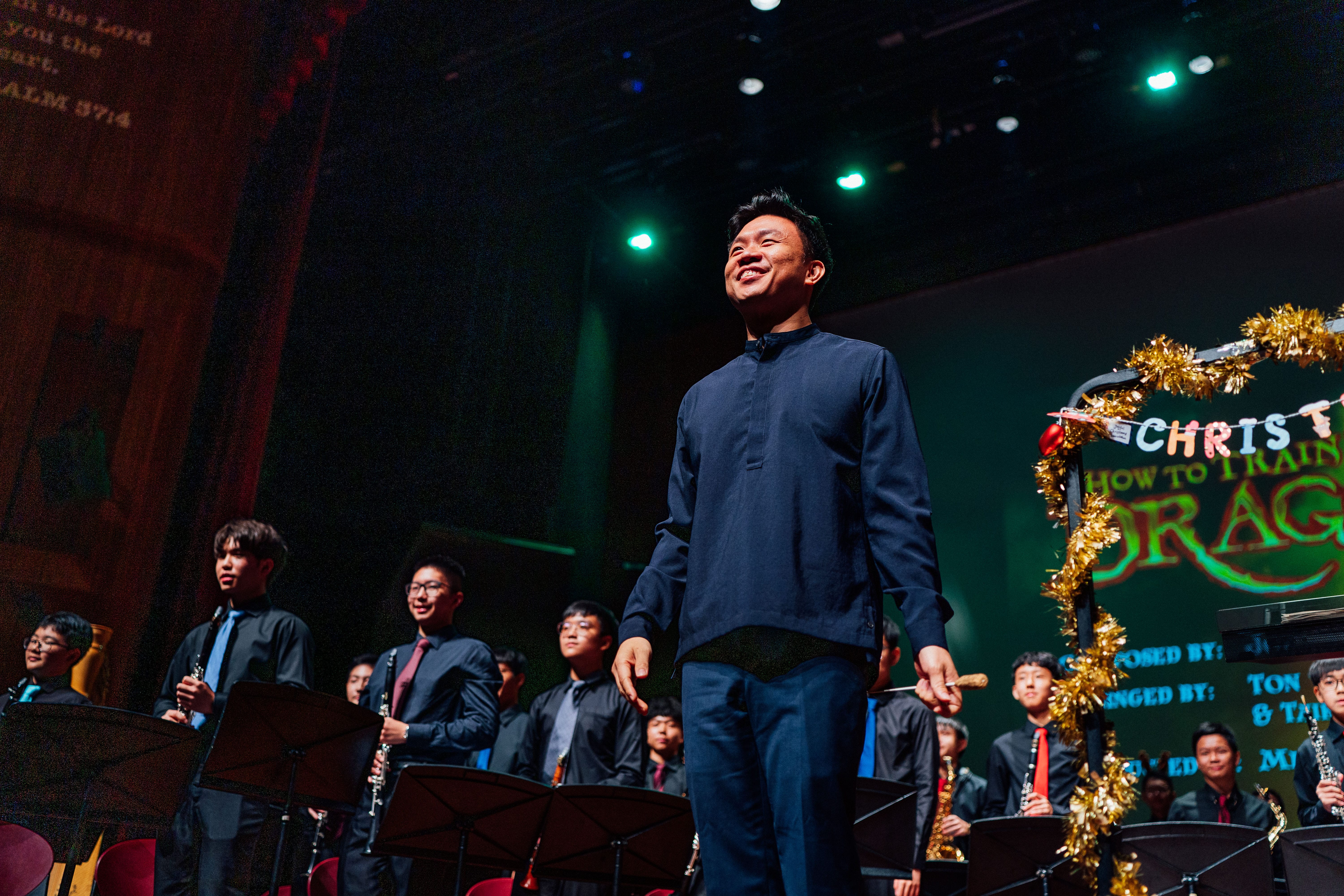 Smiling man stands on stage before a student band in black; a tinsel-framed holiday sign is behind them.