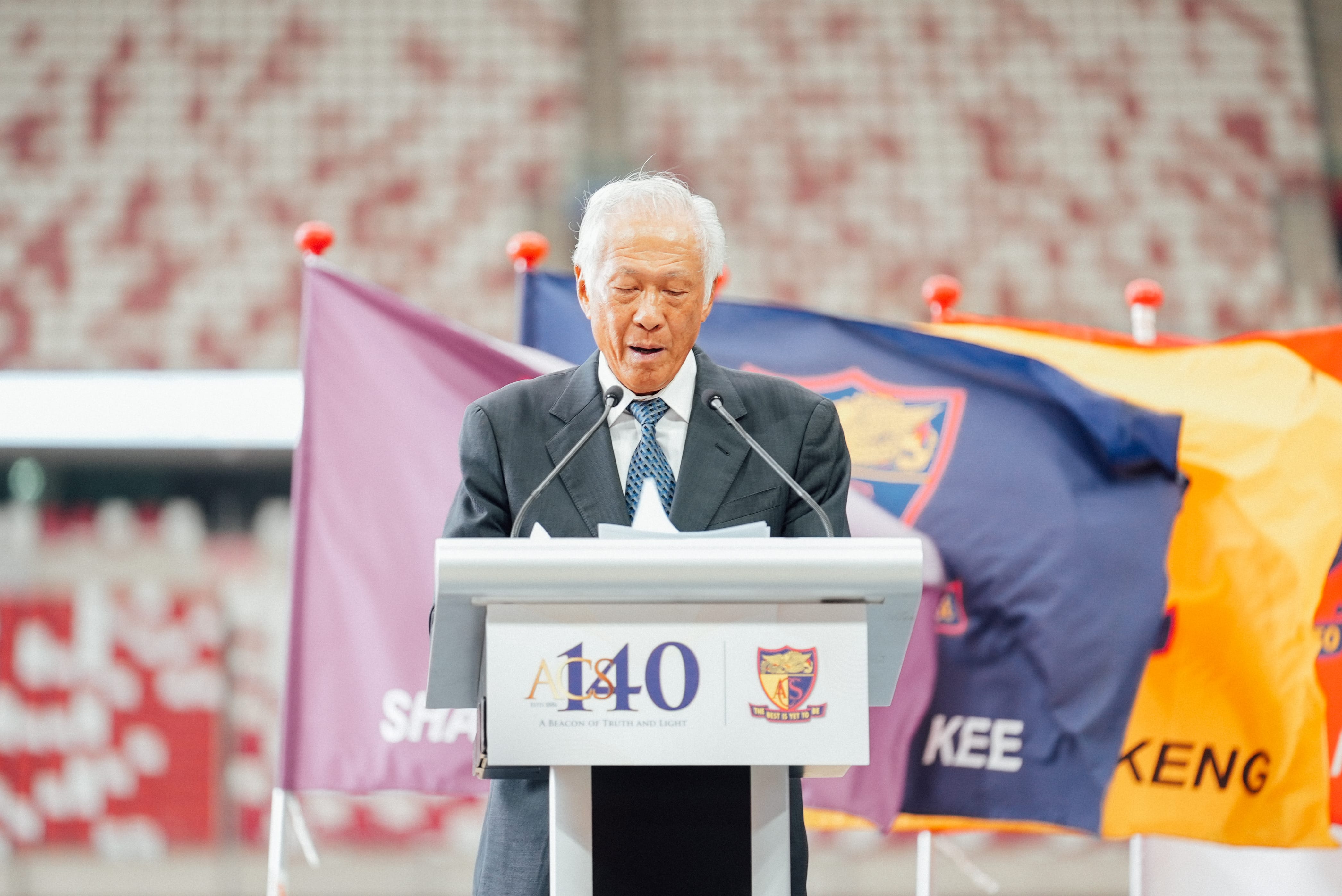 Elderly man in a suit speaking at a podium marked ACS 140, with colorful flags in the background.