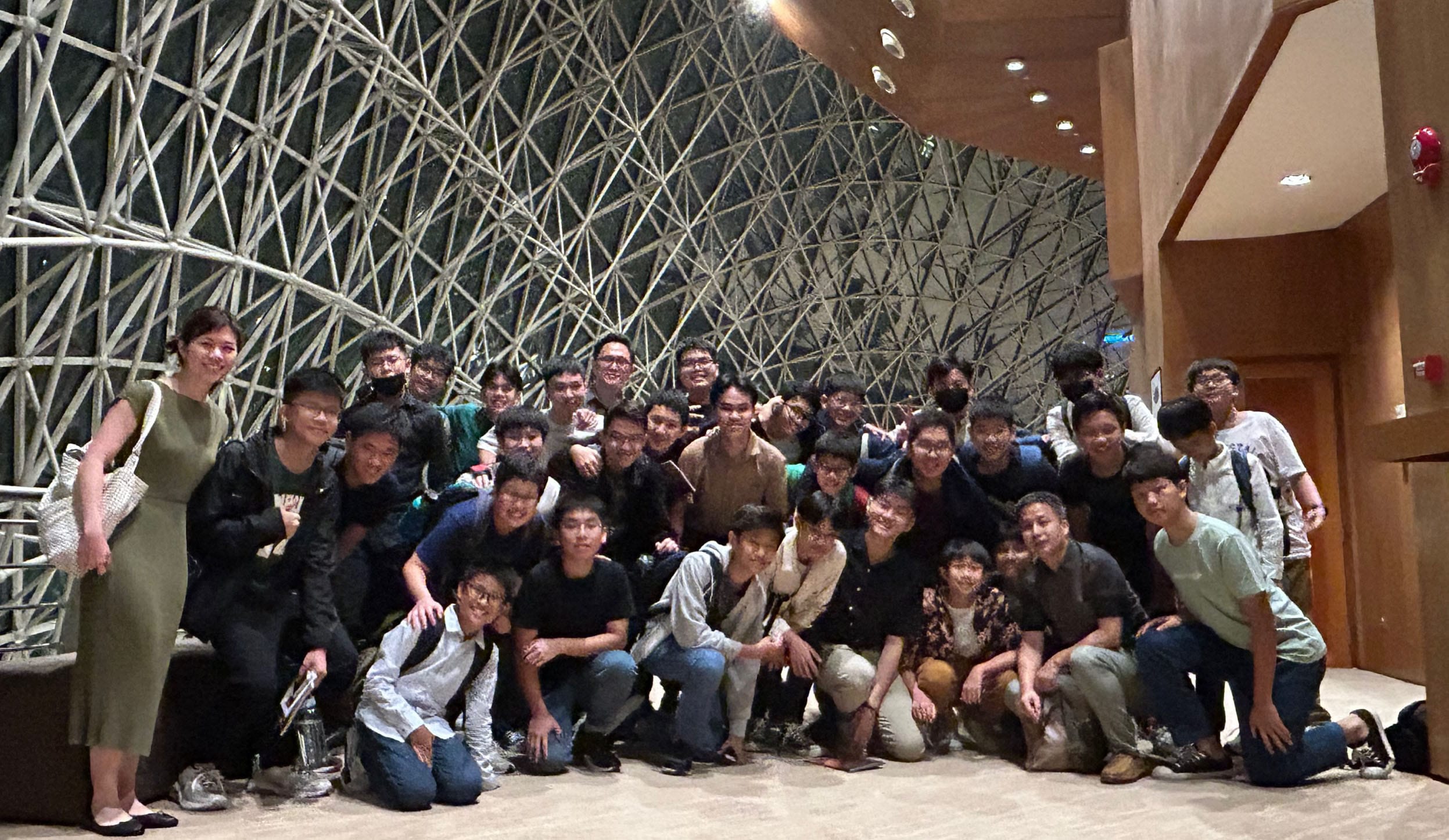 A large group of young people posing together indoors in front of a dramatic latticed wall structure.