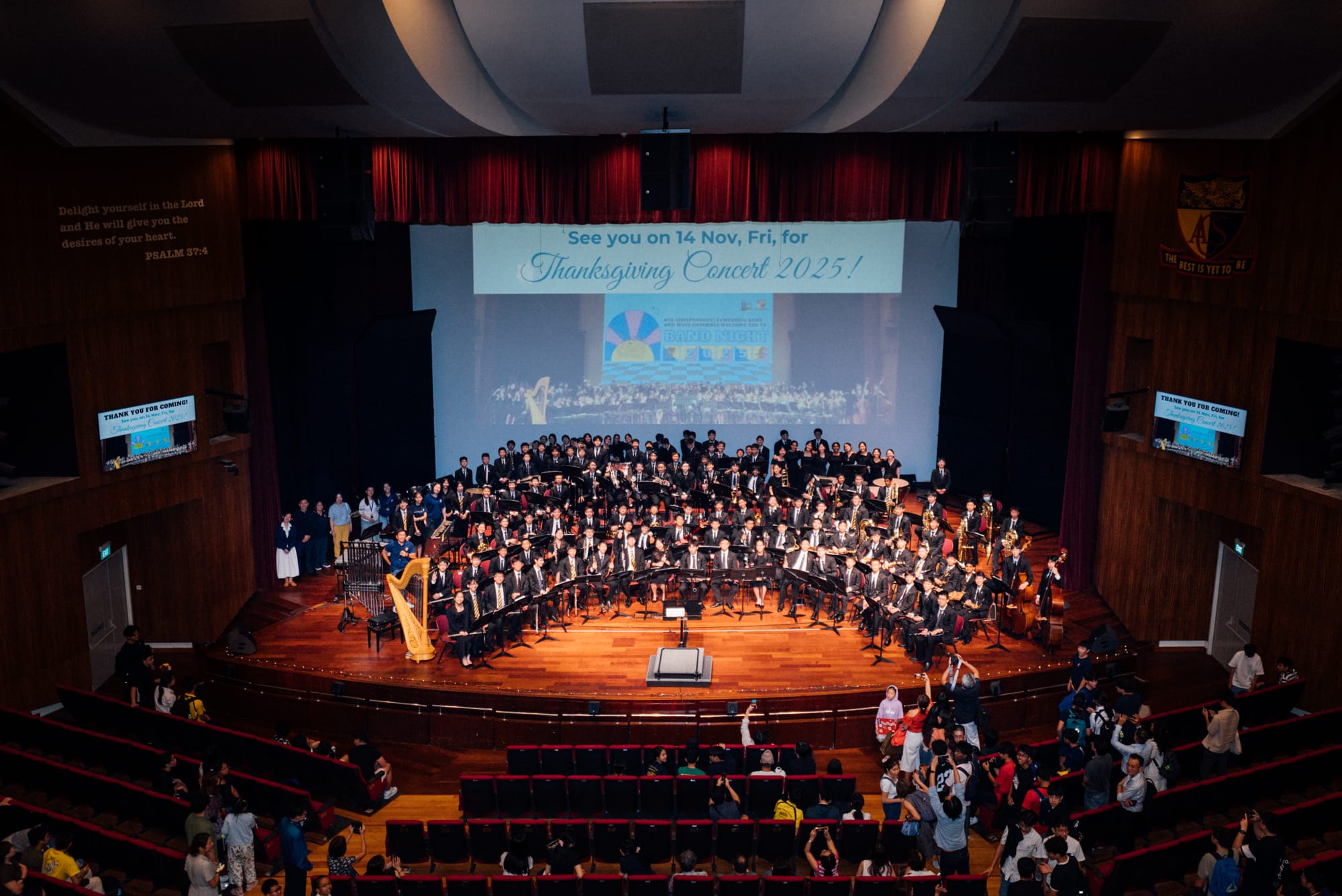 Large school orchestra on a wood-paneled stage with a projected screen reading "Thanksgiving Concert 2025" and a full auditorium.