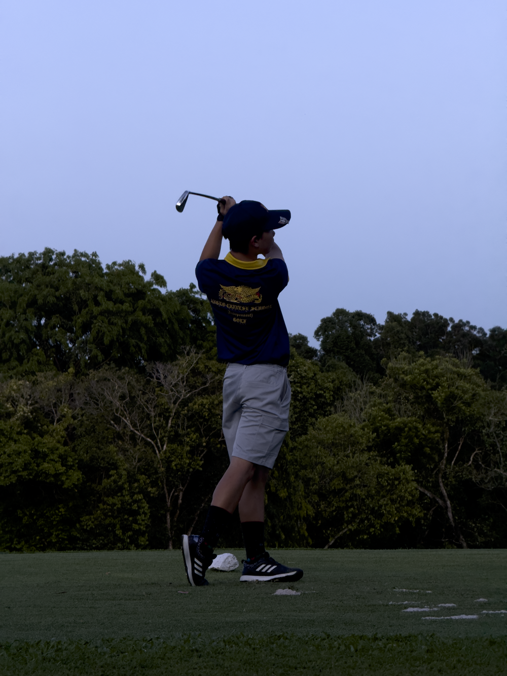 A young golfer in a blue shirt and cap follows through a swing on a grass course with trees in the background.