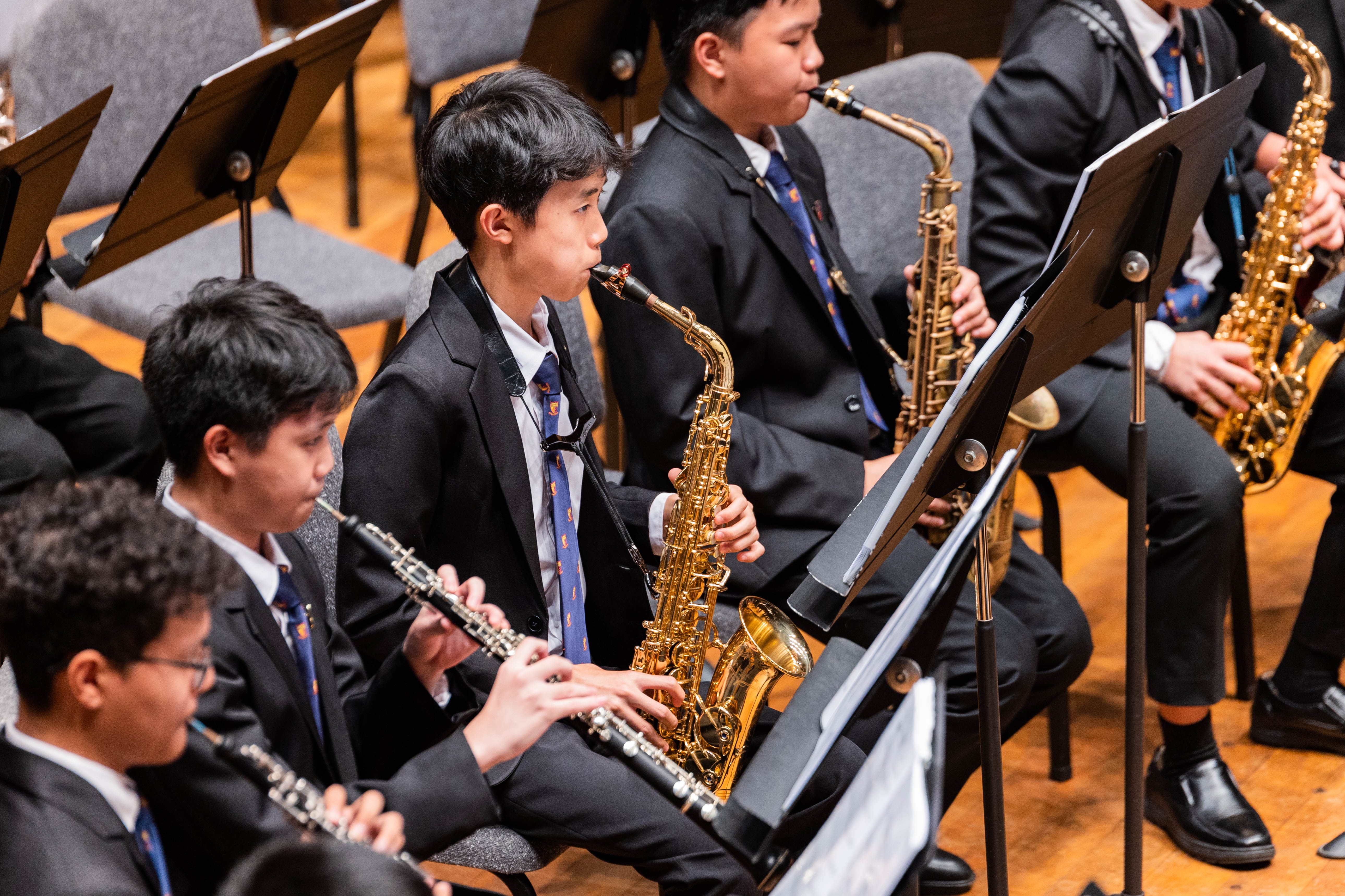 Young students in black suits and blue ties perform in a band, playing saxophones, clarinets, and flutes from sheet music.