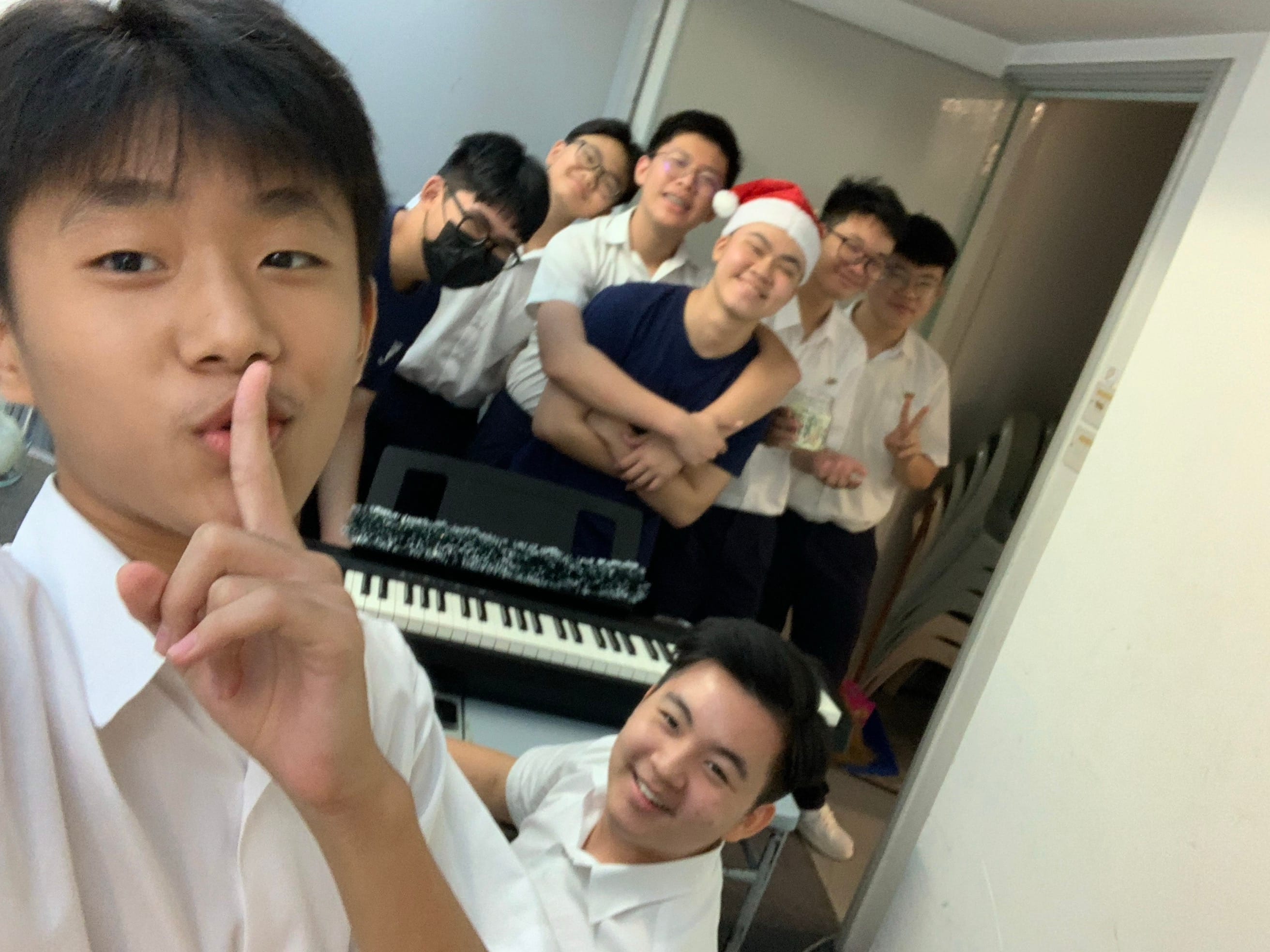 Seven young men pose in a room with a keyboard; one wears a Santa hat and another holds a finger to his lips.