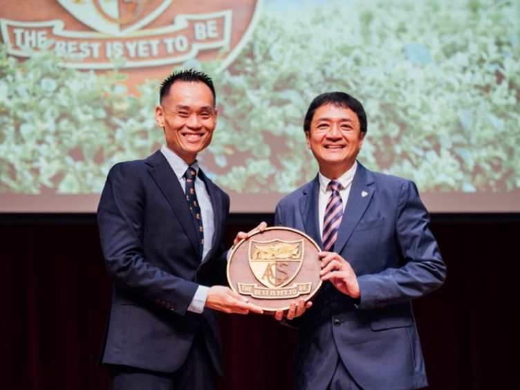 Two men in suits smile while holding a wooden shield plaque inscribed with "The Best Is Yet To Be" on stage.