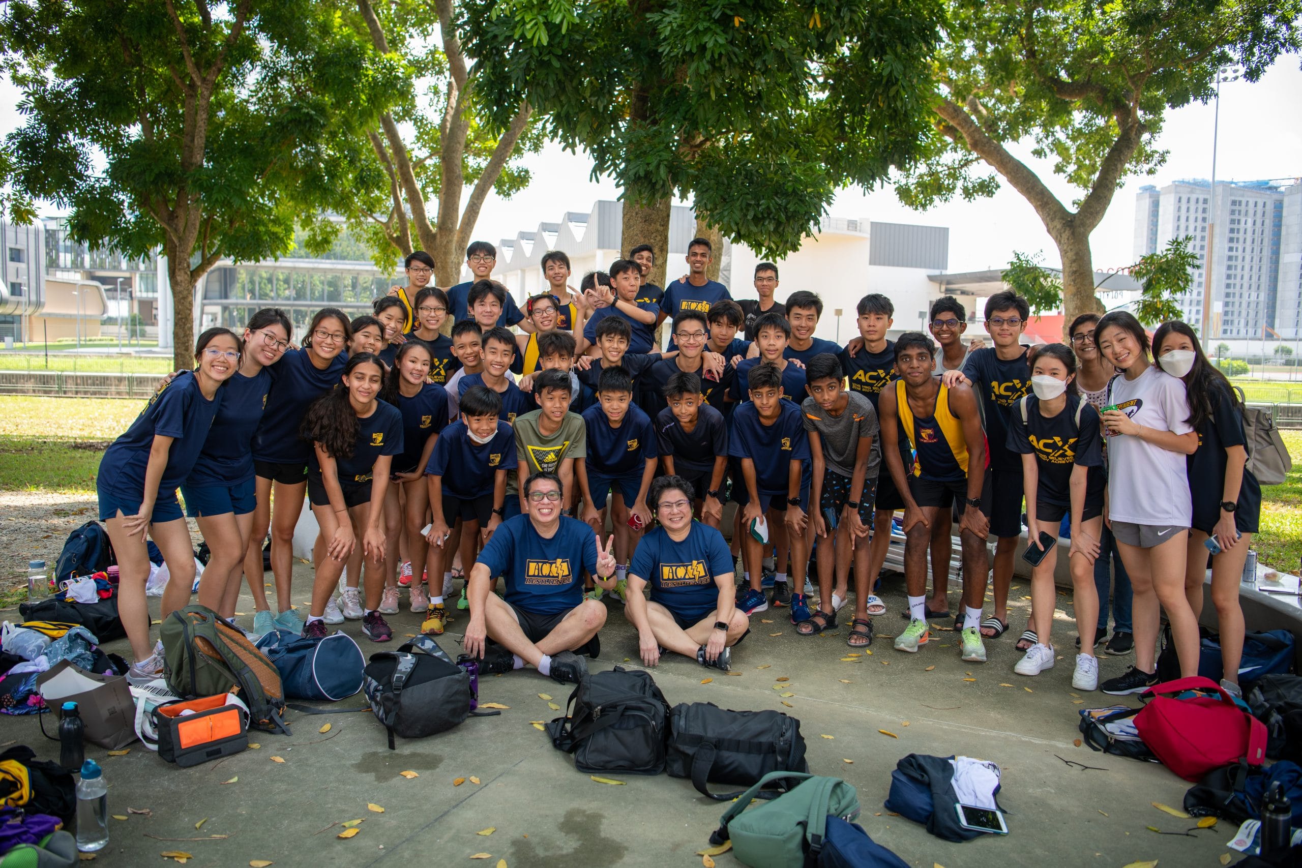 Large group of young students in matching navy blue team shirts posing outdoors under trees with bags on the ground.