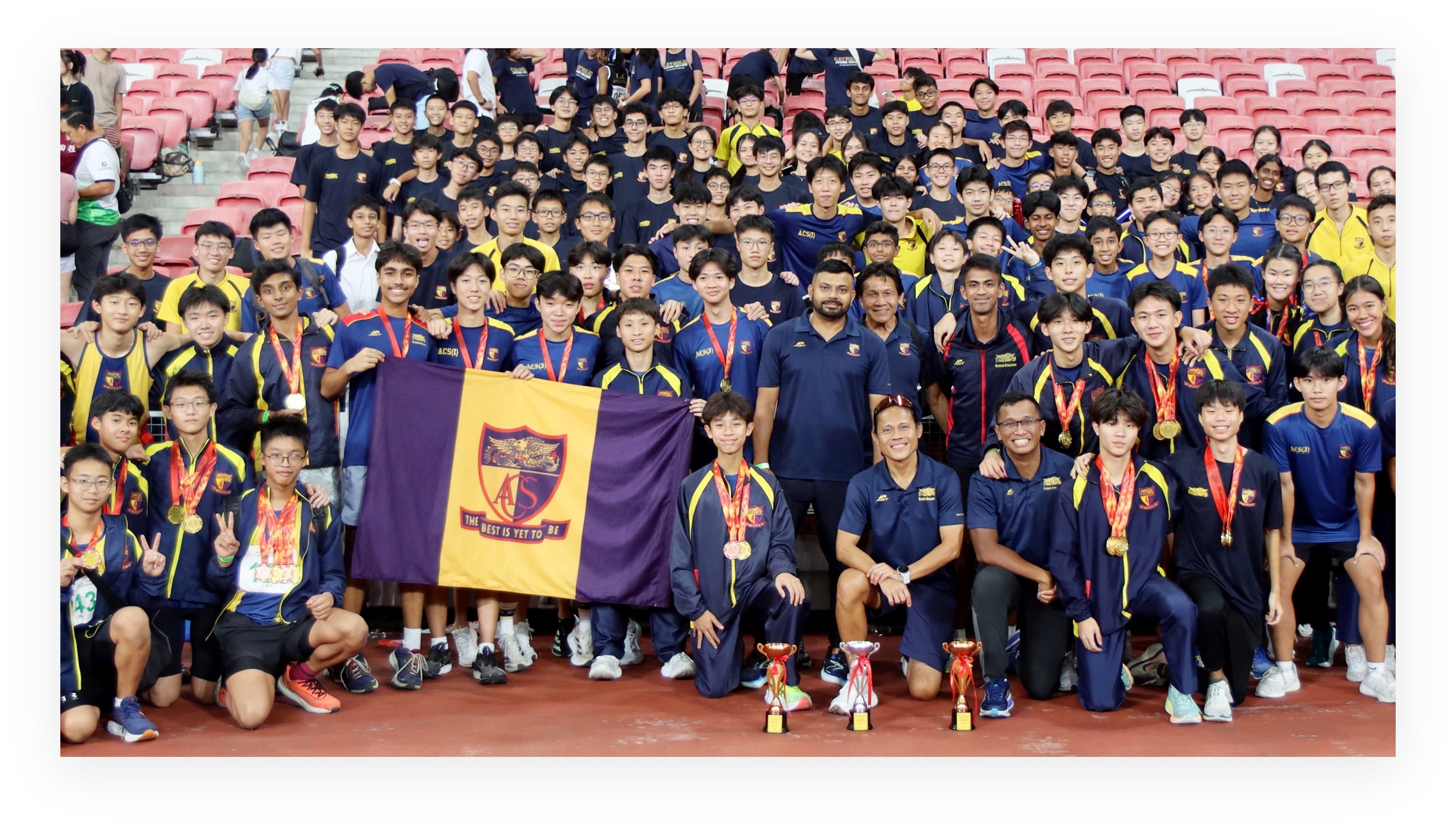 Large school team in navy and yellow uniforms poses with medals, trophies, and a purple school flag at a stadium.