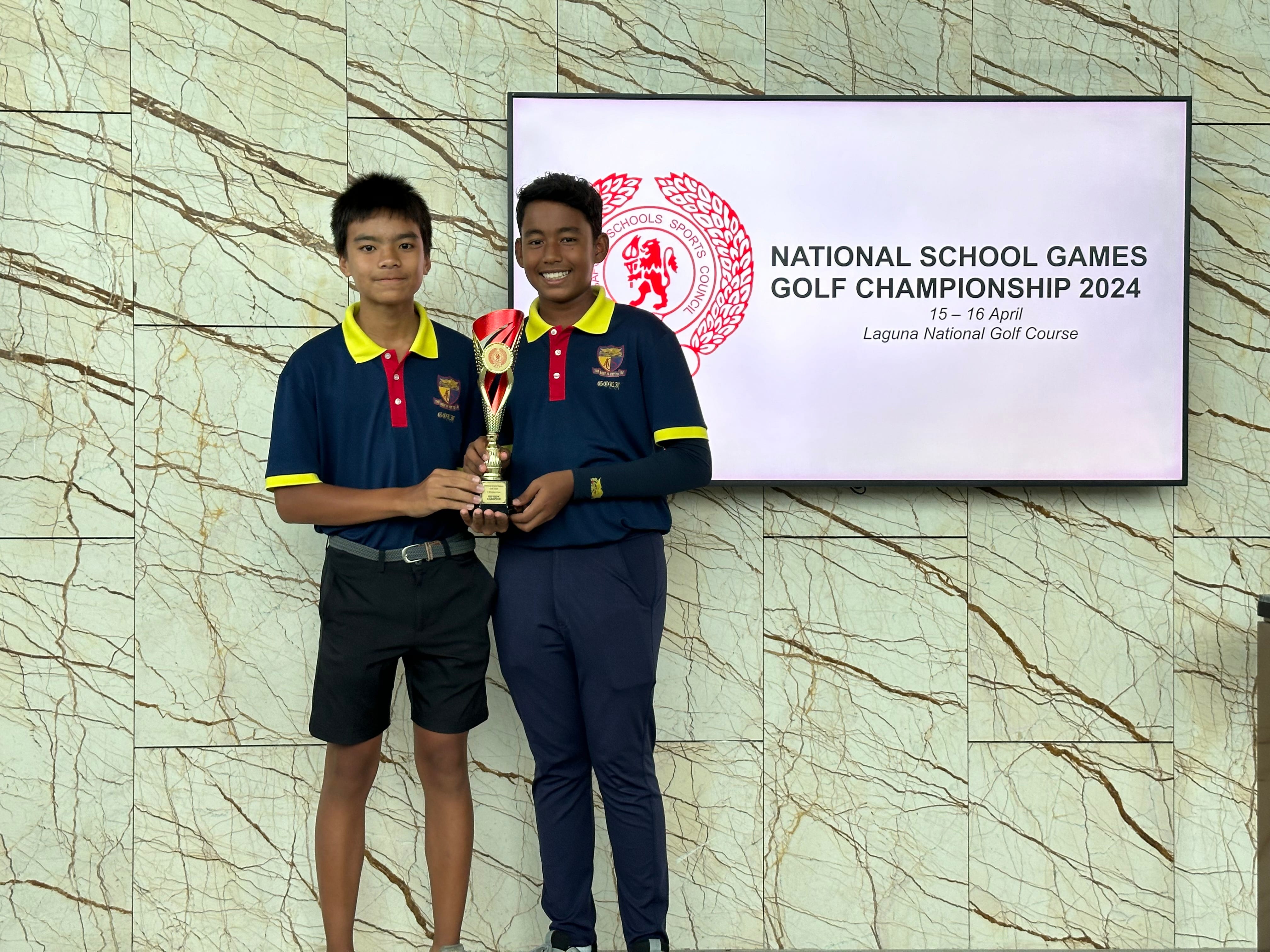 Two boys in navy and yellow polo shirts hold a trophy before a National School Games Golf Championship 2024 screen.
