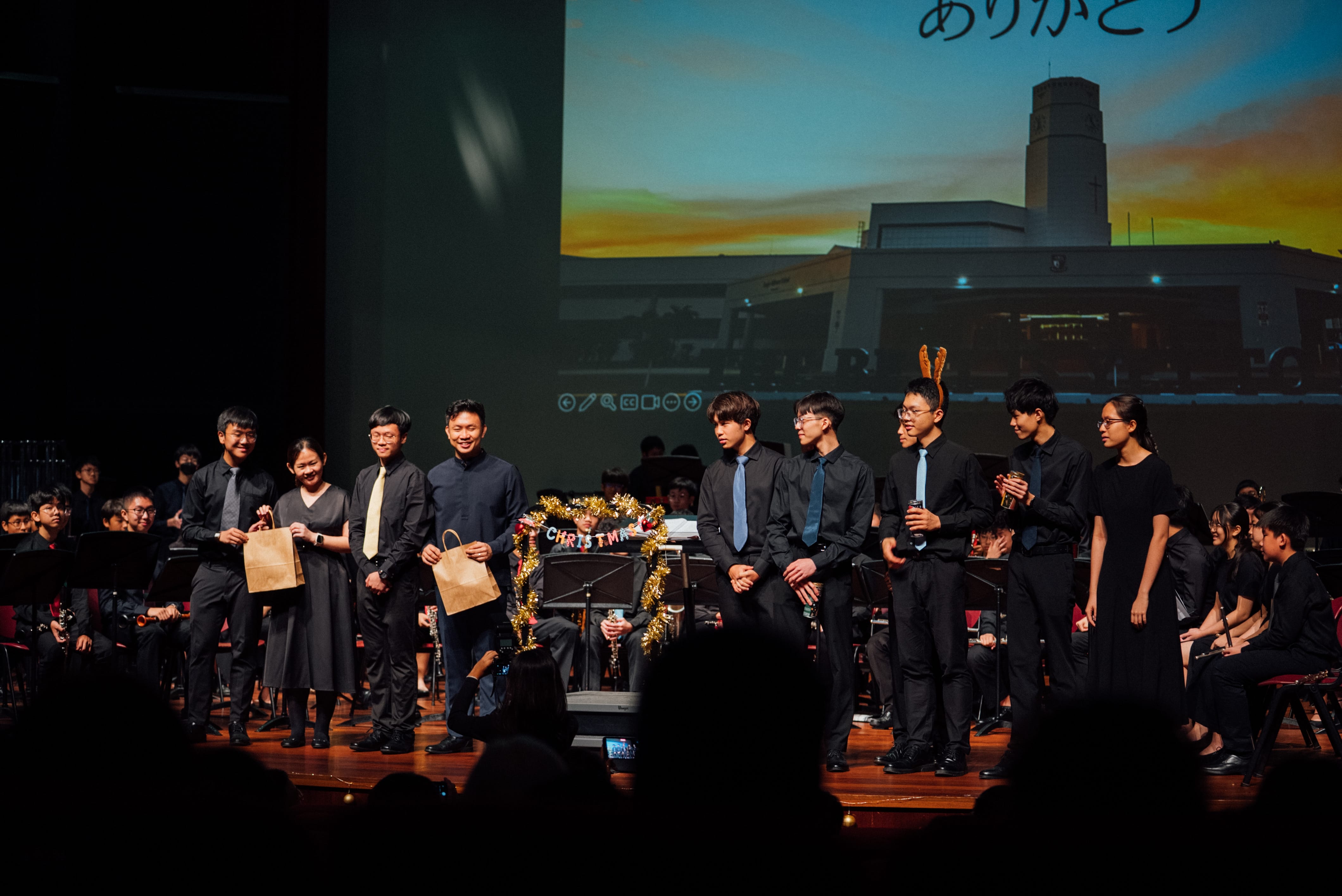 Students in black formal wear stand on a concert stage with a Christmas garland and a projected screen behind them.