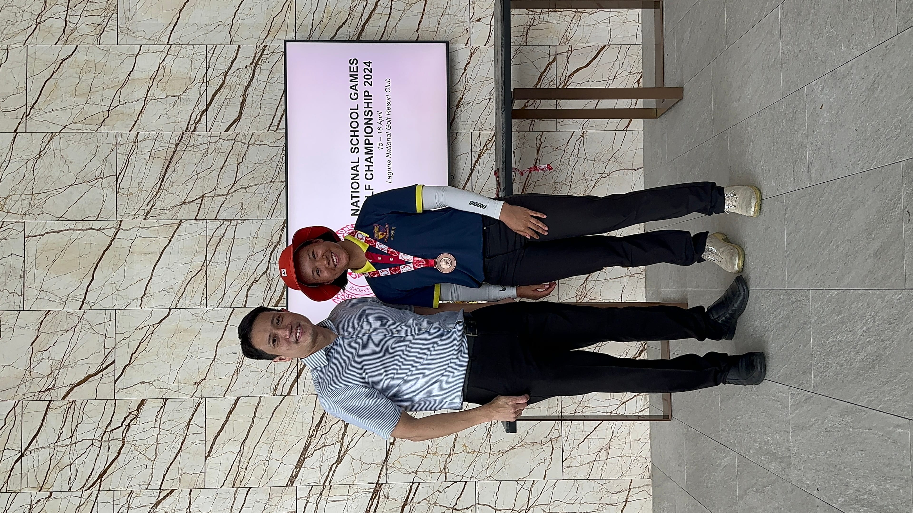 Two people pose smiling before a screen reading National School Games Golf Championship 2024, one wearing a medal.