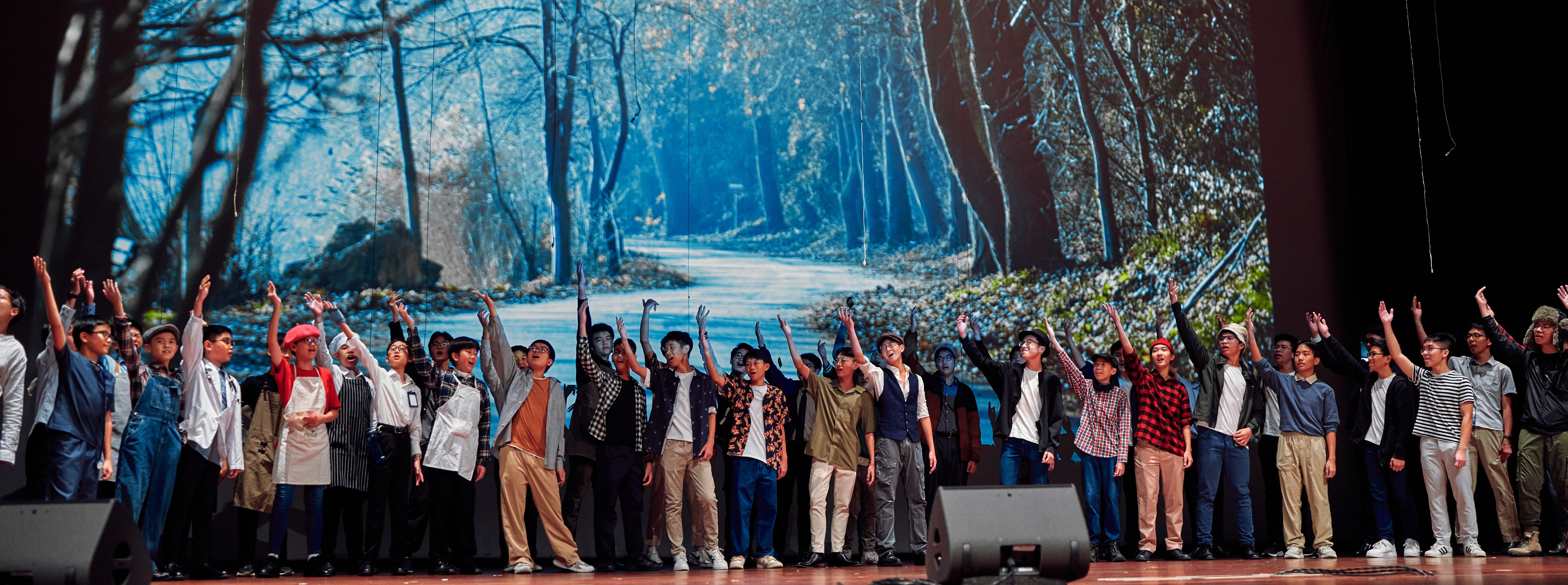 A large group of young performers on stage raising their hands, with a forest scene projected behind them.