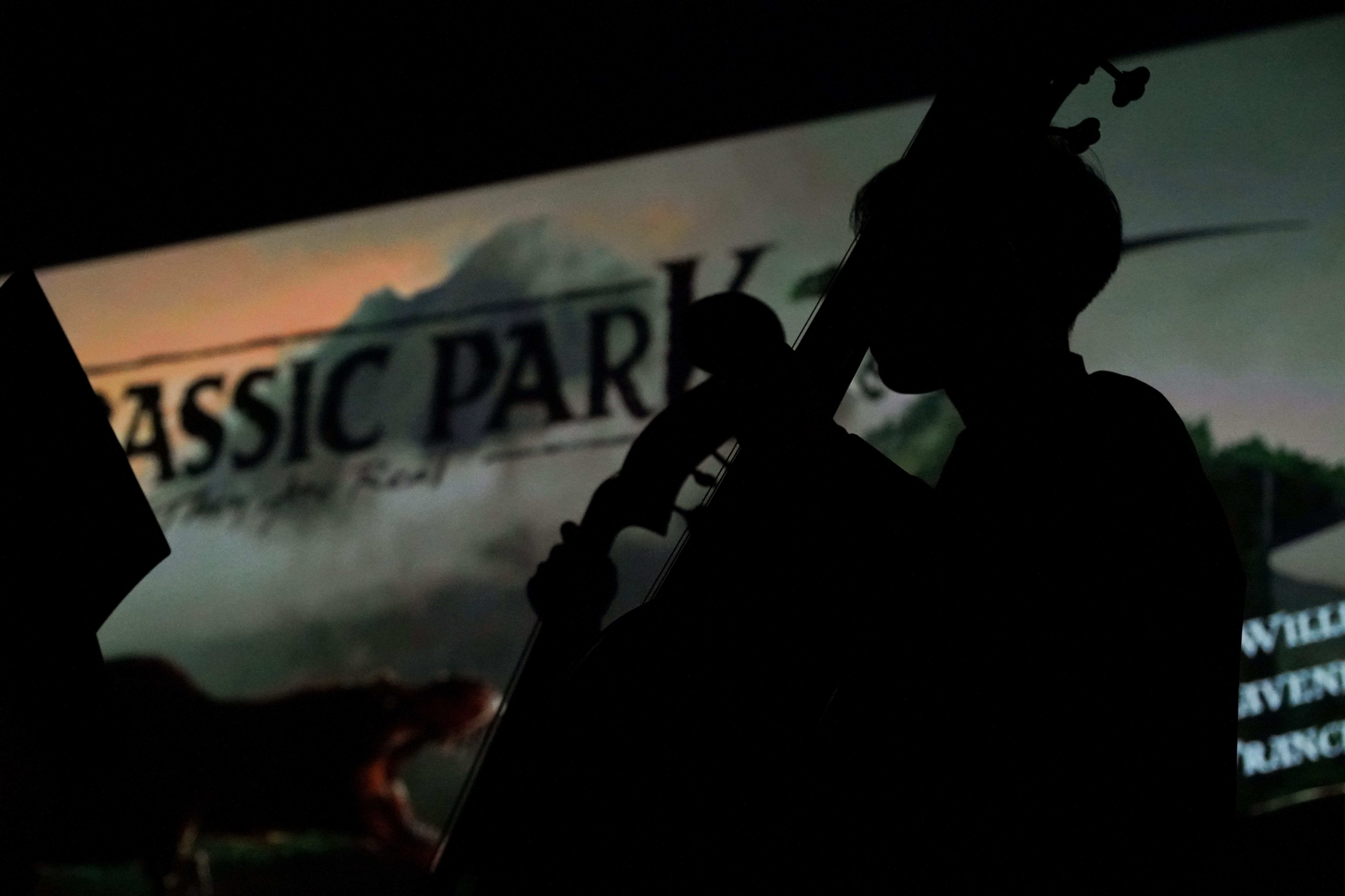 Silhouette of a cellist performing in front of a projected Jurassic Park title screen in a dark venue.